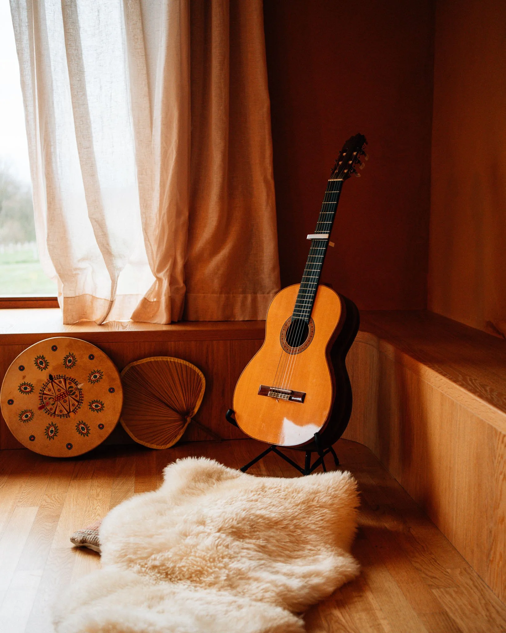 An acoustic guitar leaning on a stand in front of a window with beige curtains. To the left, a decorative round drum and an umbrella-shaped wooden piece are visible. There is a fluffy, cream-colored rug on the wooden floor, and a dark brown wall.