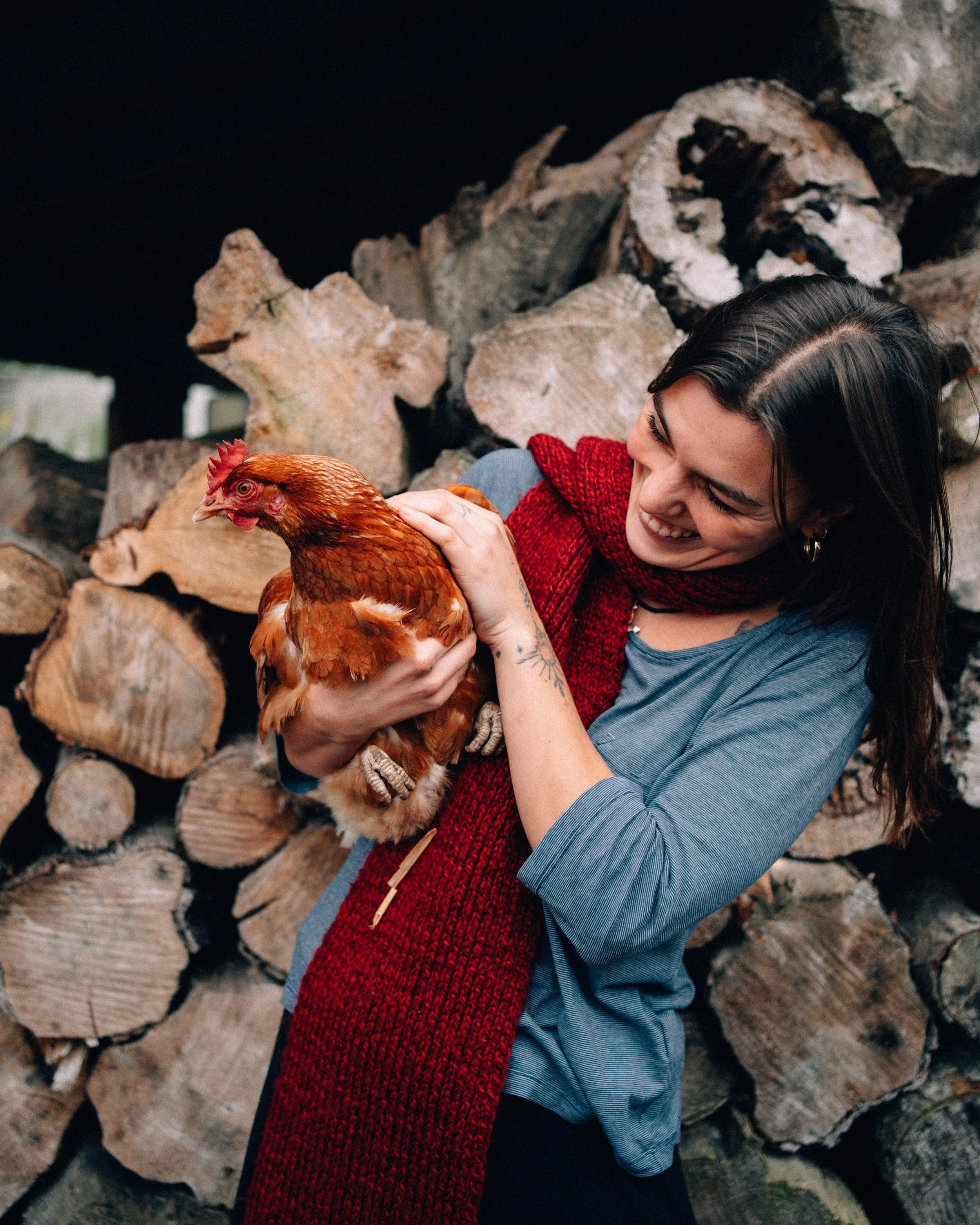 Woman with dark hair, wearing a blue shirt and red scarf, smiling and holding a brown chicken, with stacked firewood in the background.