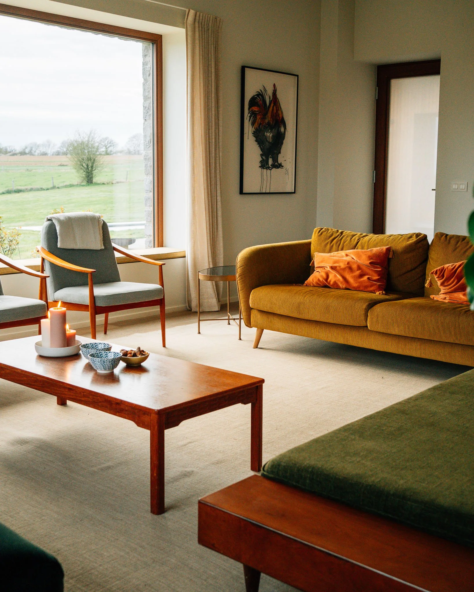 Living room with large window overlooking a green landscape, featuring a yellow sofa with orange cushions, a tan armchair near the window, a round side table, a coffee table with candles and snacks, and a colorful rooster painting on the wall.