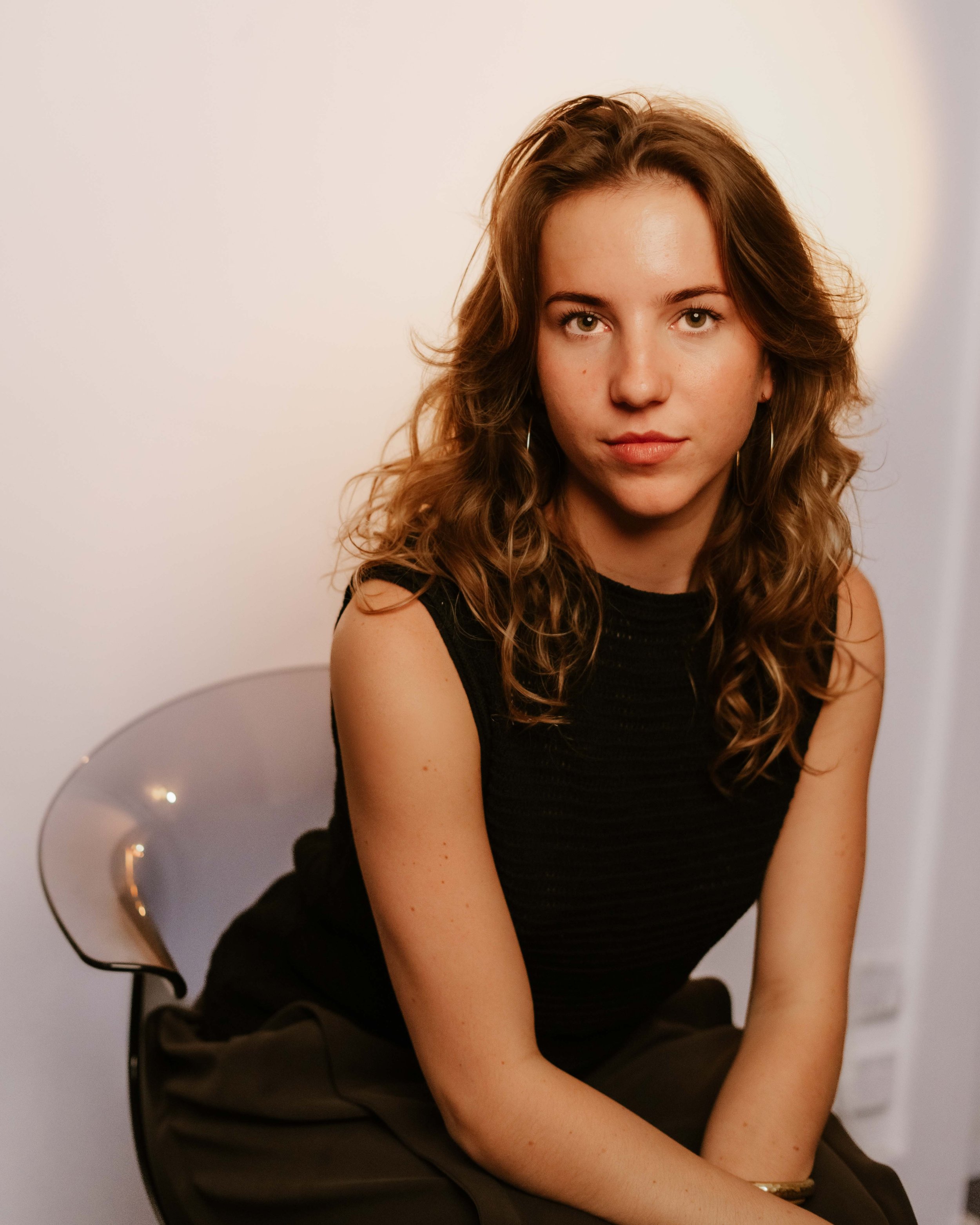 A young woman with curly brown hair and light skin sitting on a white chair with a minimalistic background. studio setting