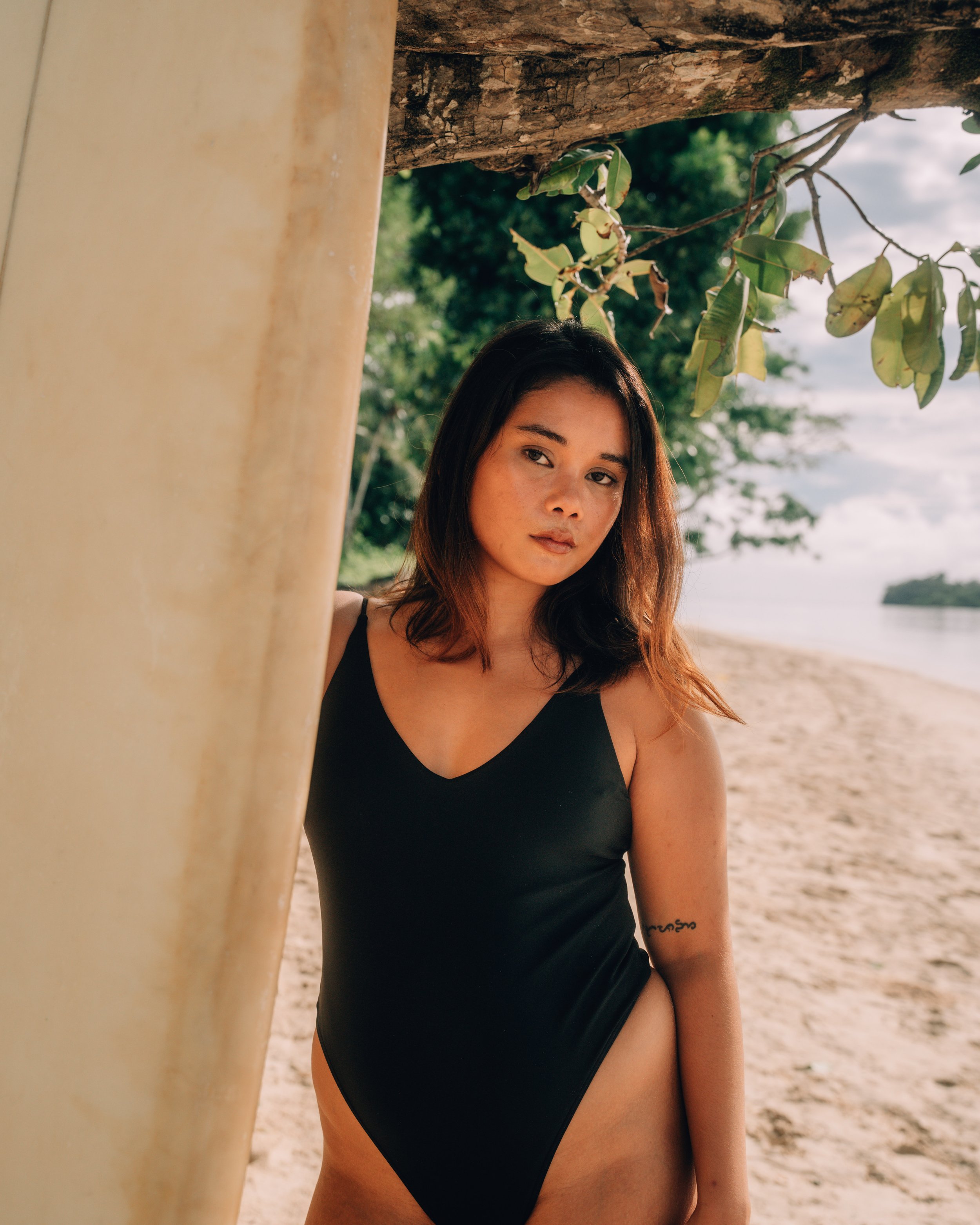 A young woman with long, wavy brown hair, wearing a black bathing suit bathingsuit, standing on a beach near a tree and a surfboard, with the ocean and an overcast sky in the background.