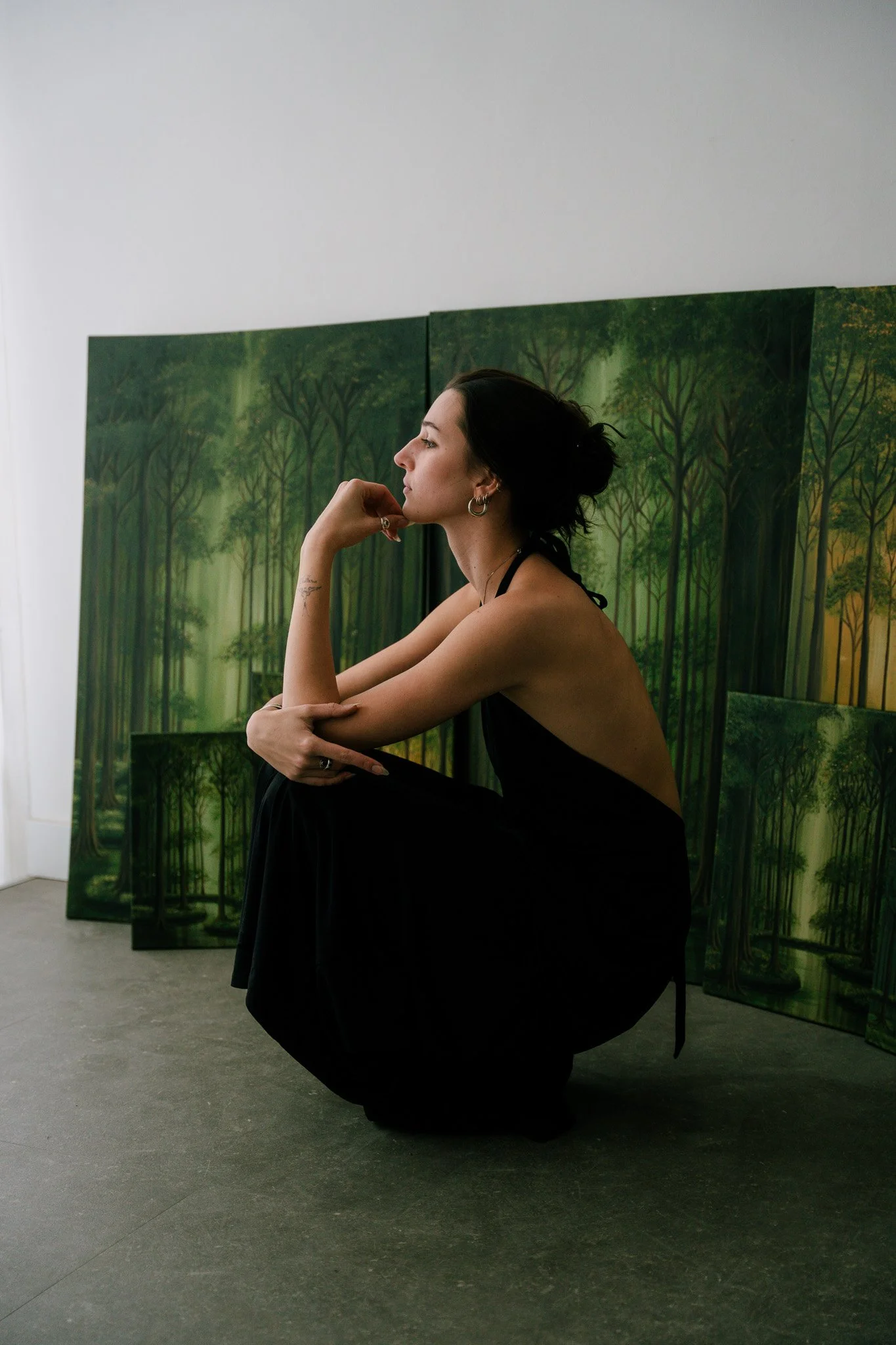 A woman crouching in front of green paintings of trees in an art gallery, resting her chin on her hand while wearing a black dress, silver hoop earrings, and multiple rings.