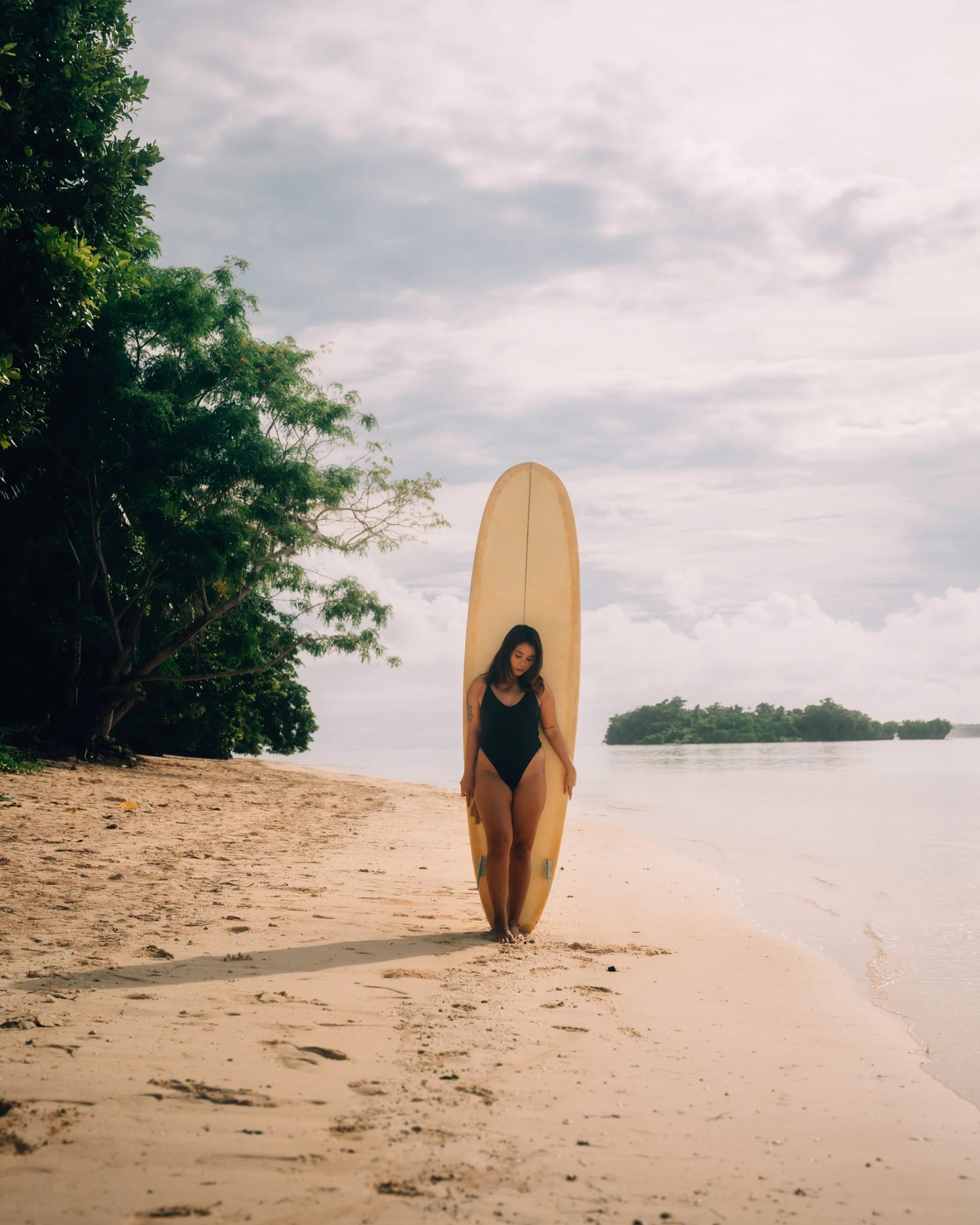 A woman in a black swimsuit holding a surfboard on a sandy beach with water and trees in the background, under a cloudy sky.