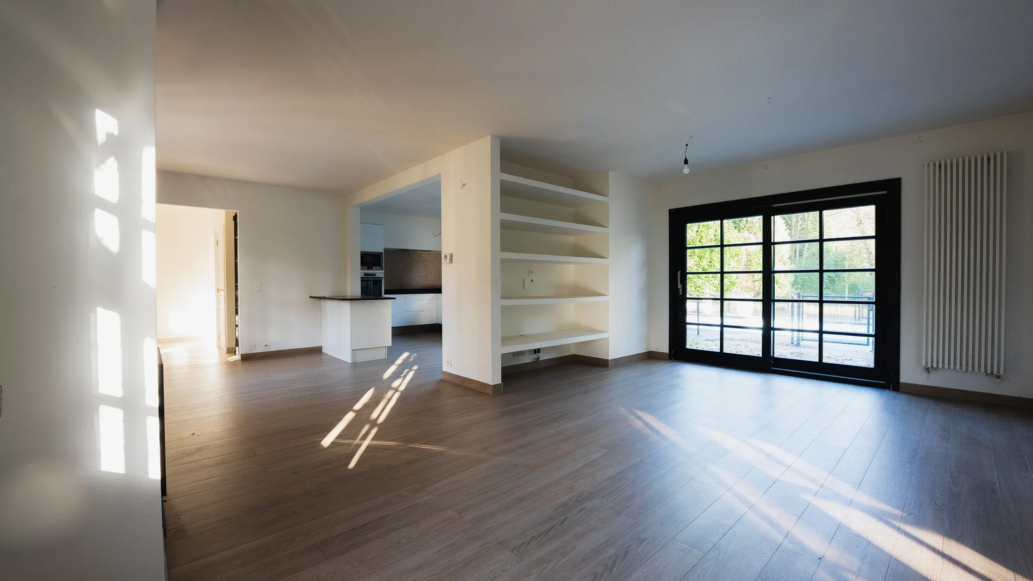 Empty living room with large black sliding glass doors, built-in shelves, and sunlight casting shadows on the wooden floor.