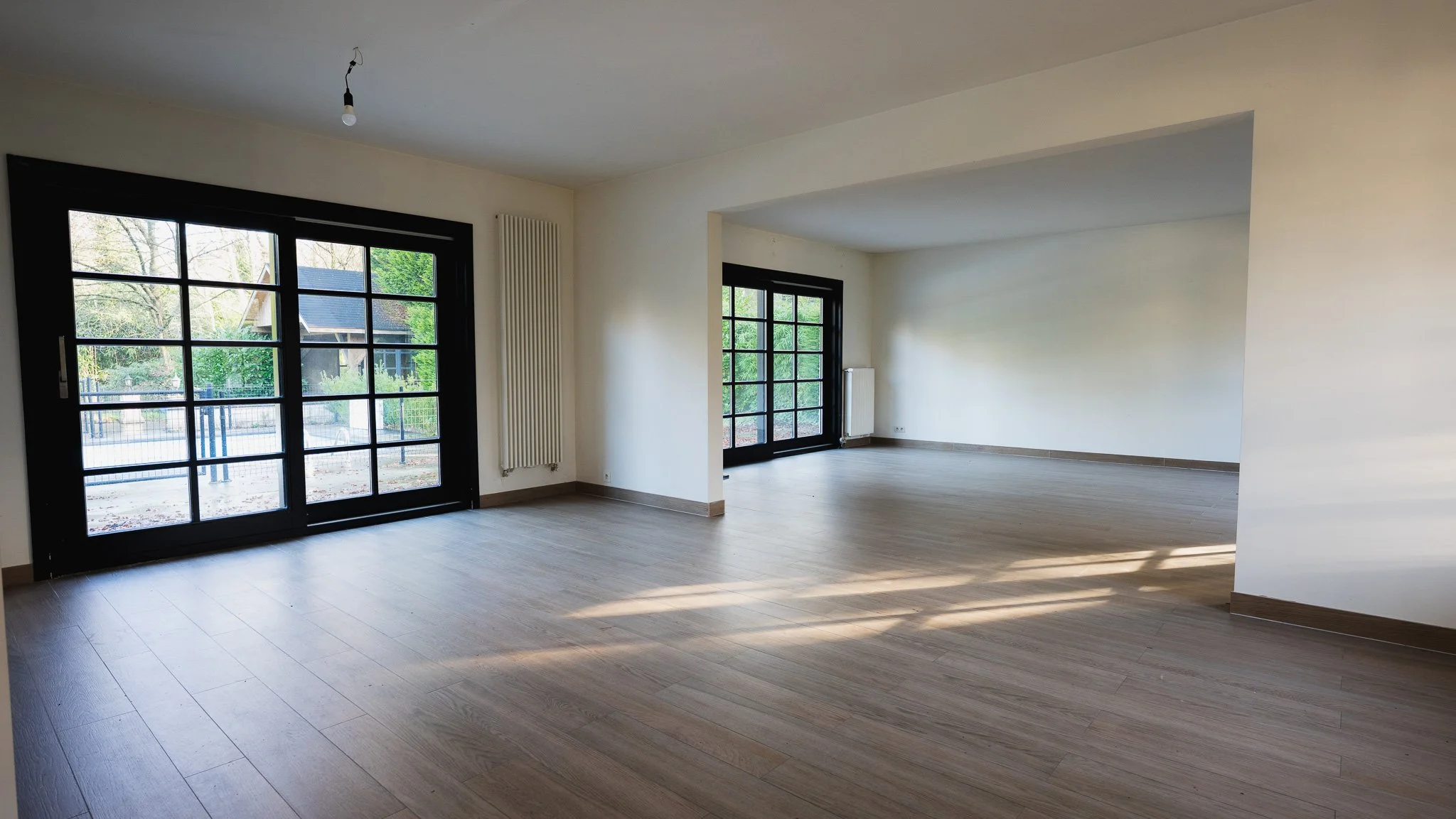 Empty living room with large black-framed glass doors leading outside, light wood flooring, white walls, and a ceiling with an exposed light bulb.
