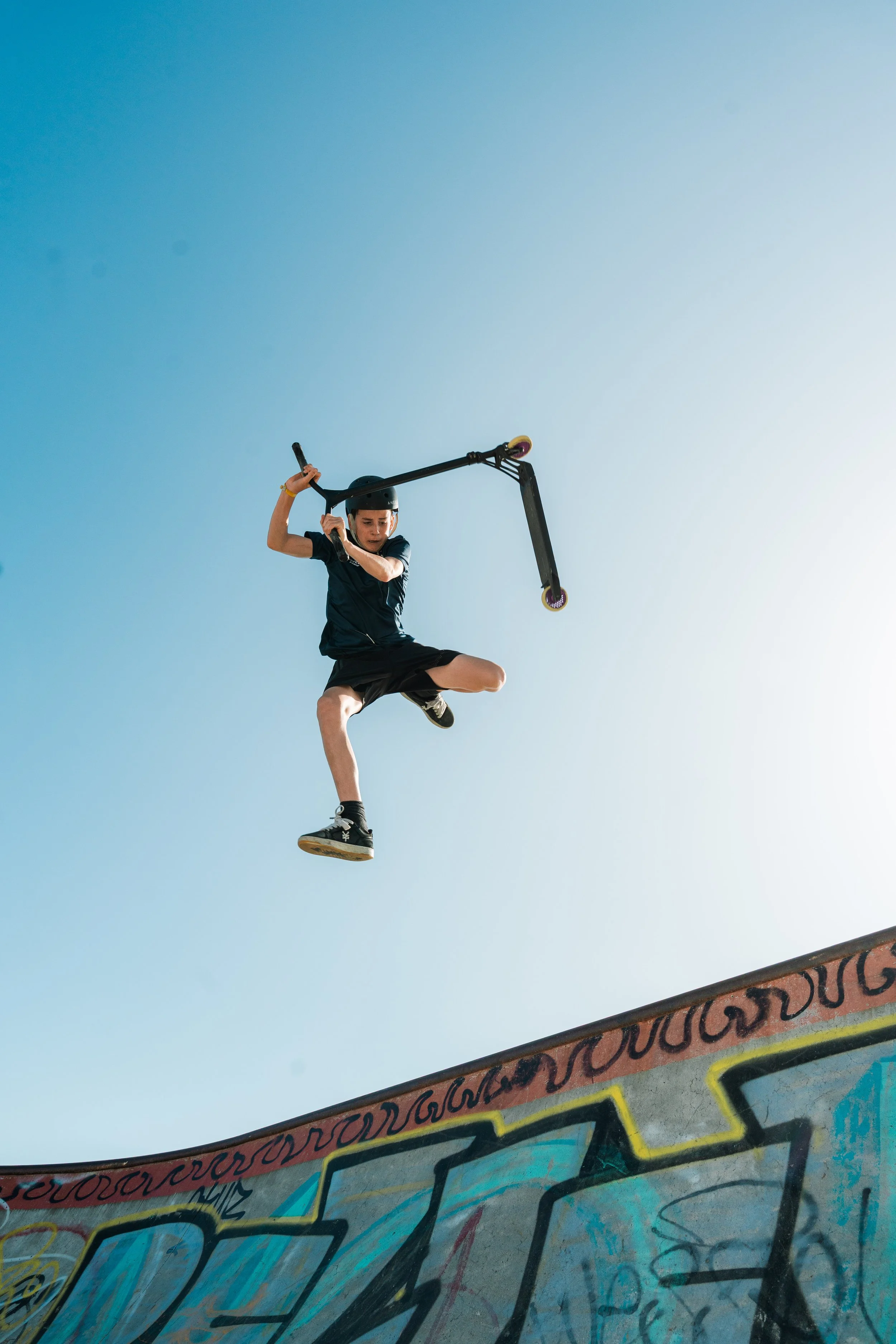 A young person wearing a helmet is mid-air on a scooter at a skate park, with a clear blue sky in the background and graffiti on the ramp below.