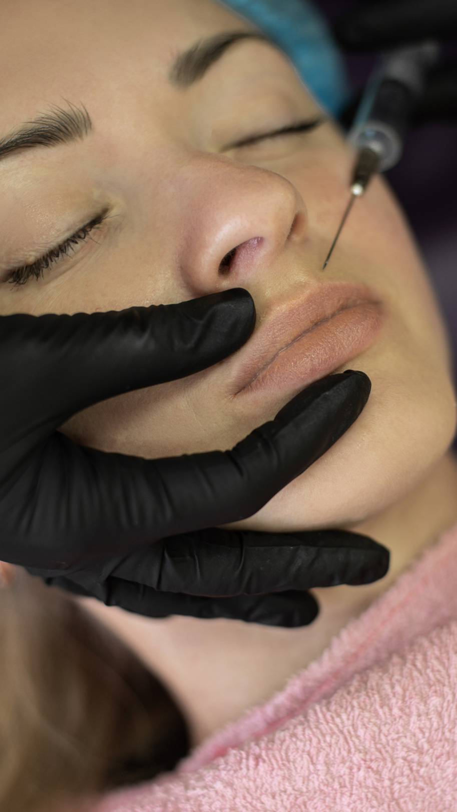A person receiving a cosmetic injection in the nose with a fine needle while lying down with eyes closed, wearing a pink towel.