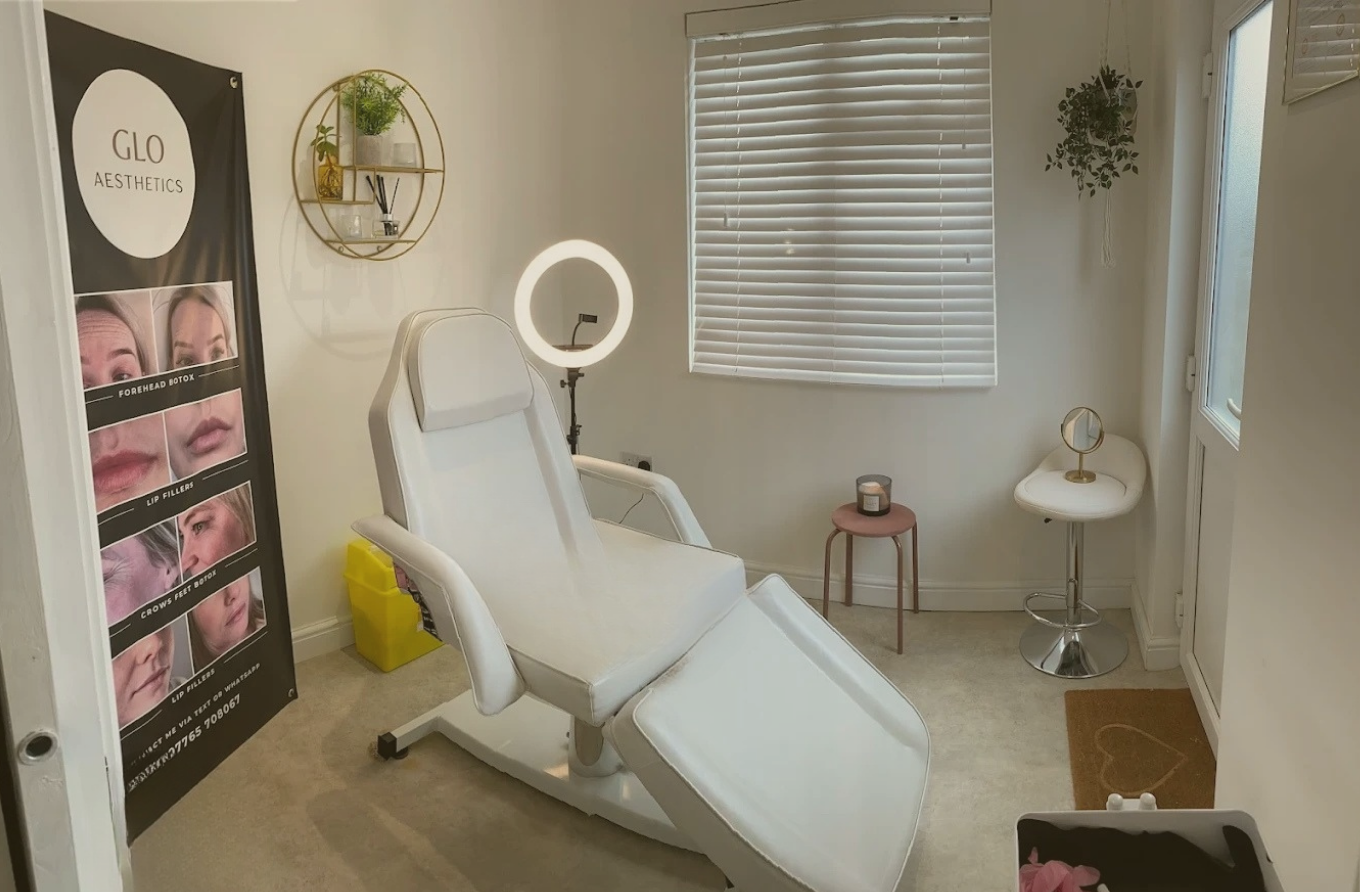 A clean, well-lit treatment room with a white adjustable chair, a ring light, a small side table with a candle, and a mirror. There is a framed advertisement on the wall showing before and after images of facial treatments. The room has blinds on the window and decorative wall shelves with plants.