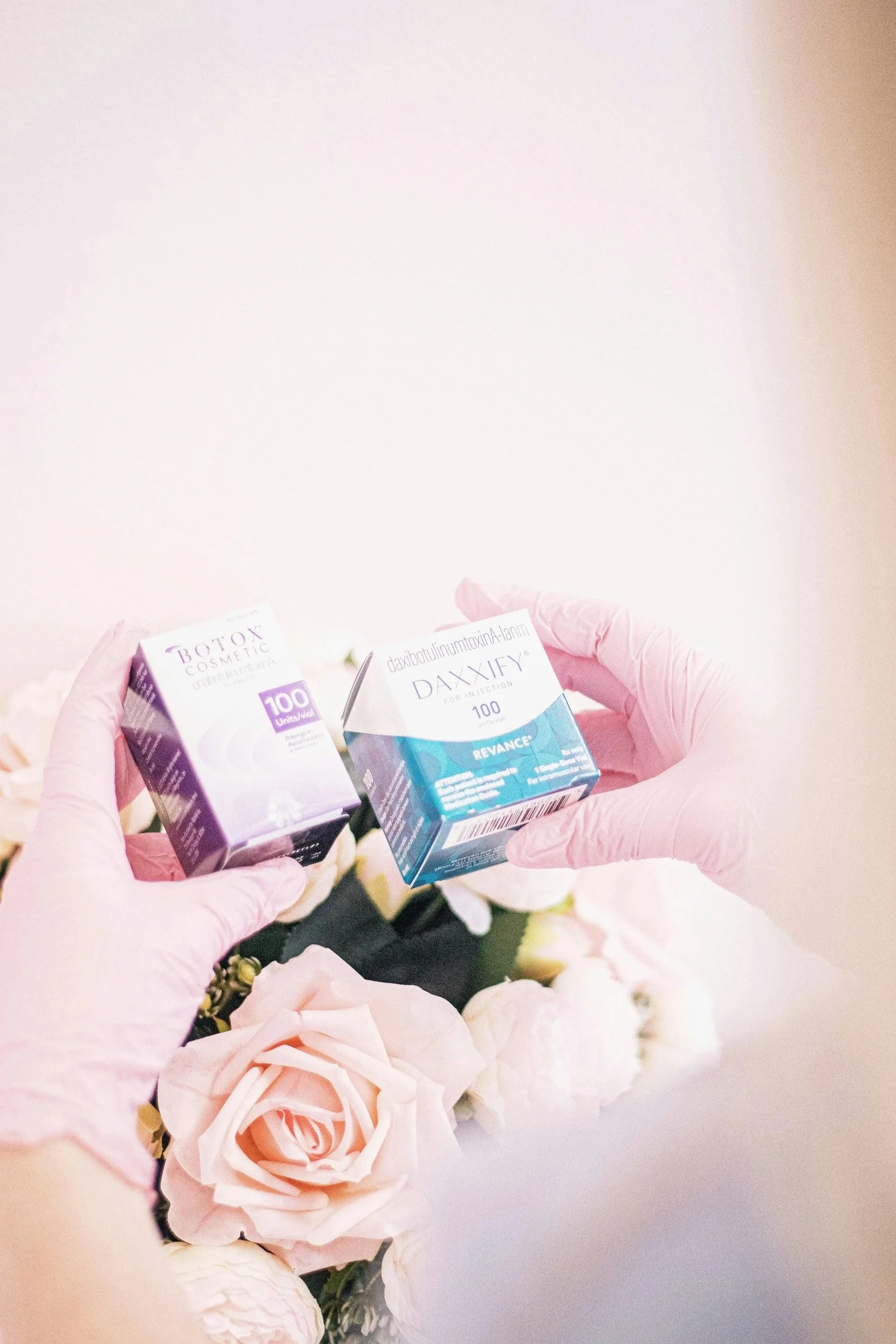Hands wearing pink gloves holding three boxes of Botox and Daxify medications in front of light pink and white roses.