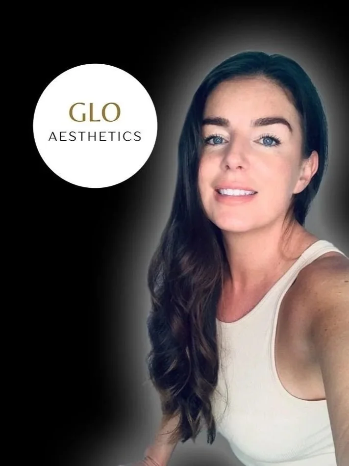 A woman with long dark hair and blue eyes taking a selfie, wearing a white sleeveless top, with a black background and a white circular logo that says "GLO AESTHETICS".