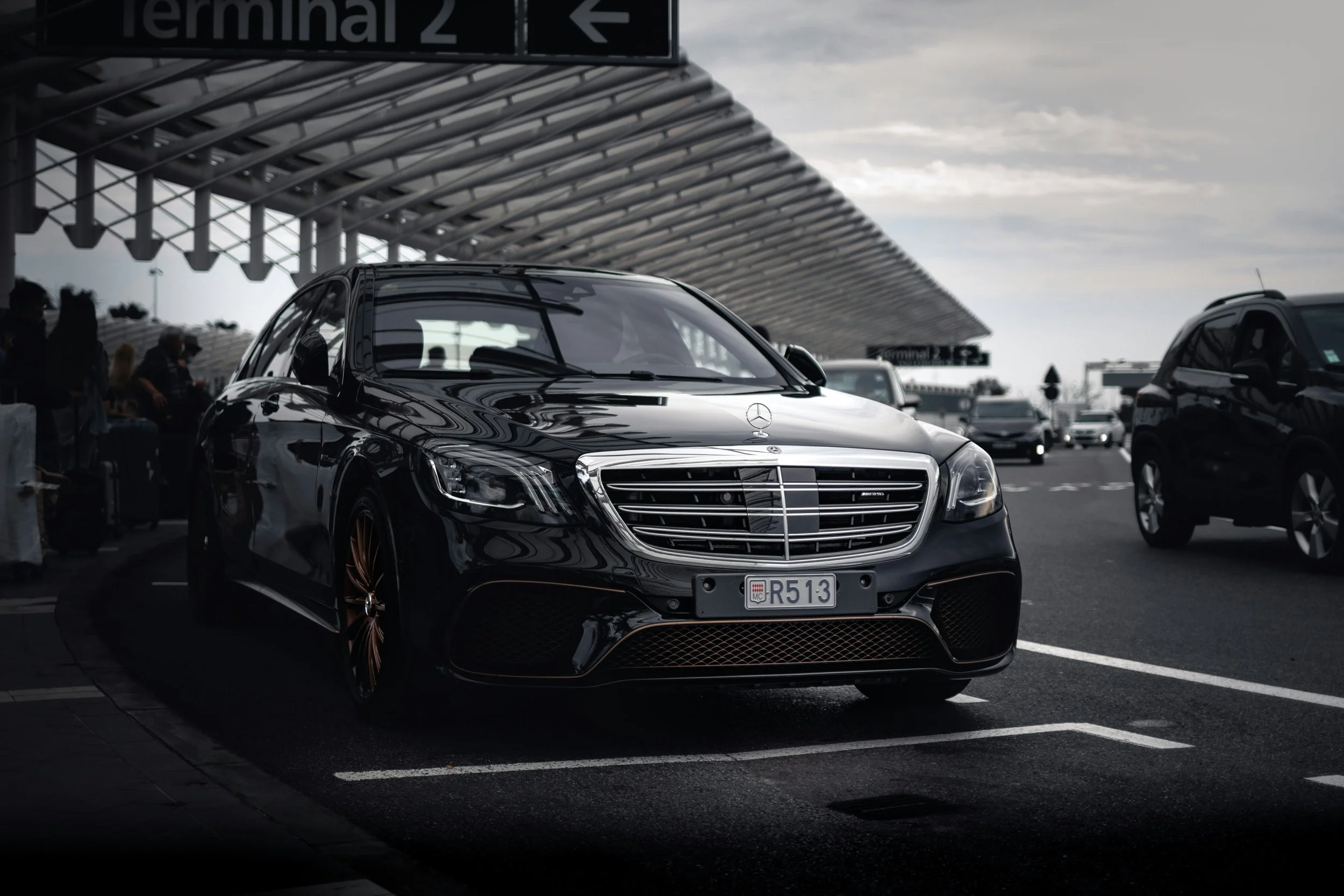 Une berline noir Mercedes-Benz stationnée dans un parking à l'extérieur, avec d'autres véhicules visibles et une structure en toiture au-dessus, sous un ciel nuageux.