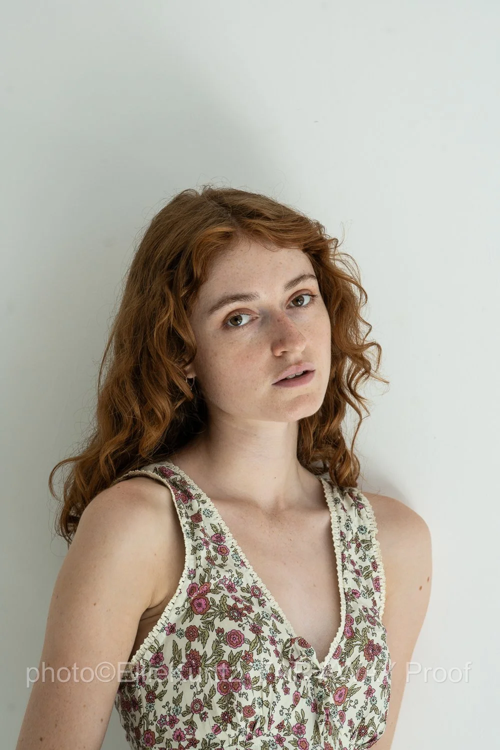 A young woman with curly red hair and freckles, wearing a floral sleeveless top, standing against a plain, light-colored background.
