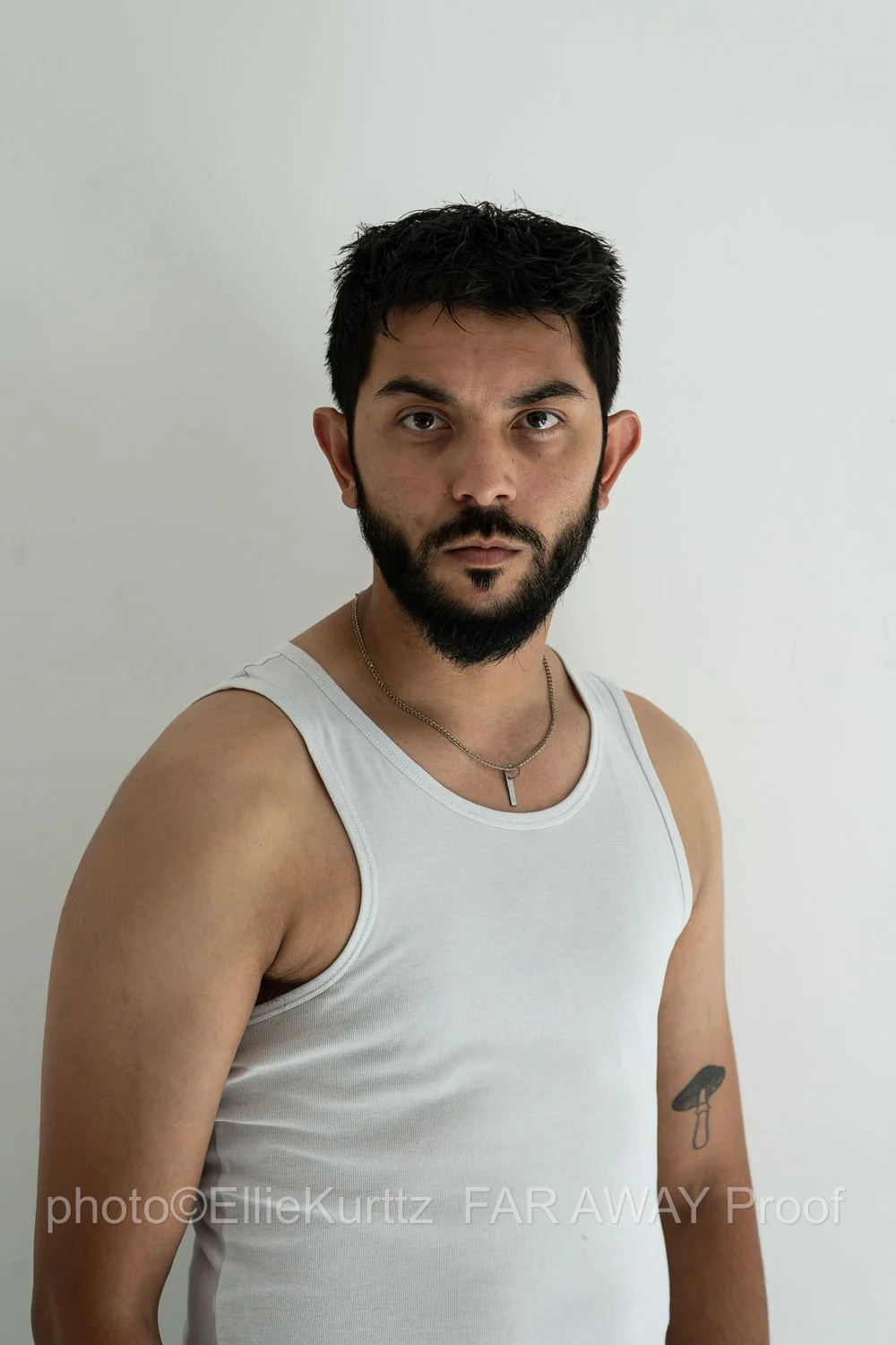 A young man with dark hair and a beard, wearing a white tank top, silver necklace, and a tattoo of a mushroom on his left arm, standing against a plain white background.
