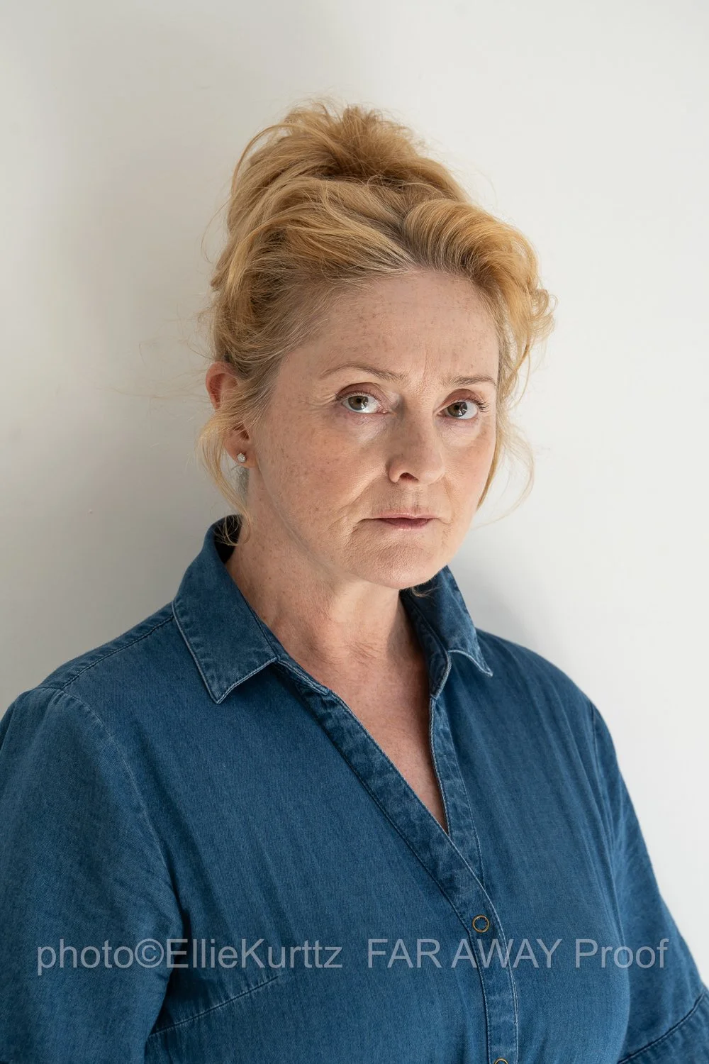 A middle-aged woman with red hair styled in an updo, wearing a blue denim shirt, standing against a plain white background.