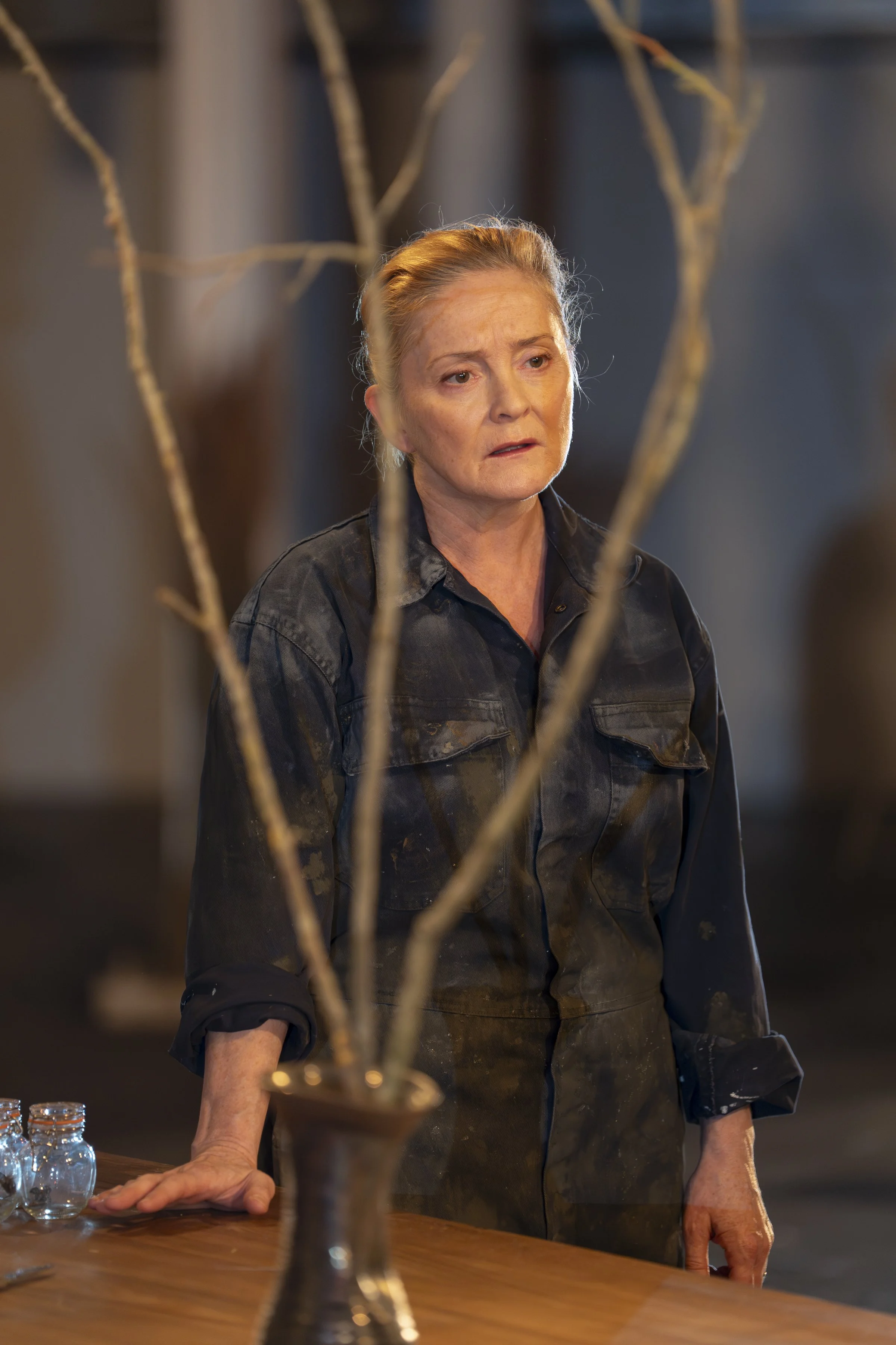A middle-aged woman with light hair tied back, wearing a black shirt and apron, standing behind a wooden table, with a decorative branch in the foreground. The woman's facial expression looks contemplative or concerned, and the background is blurred 