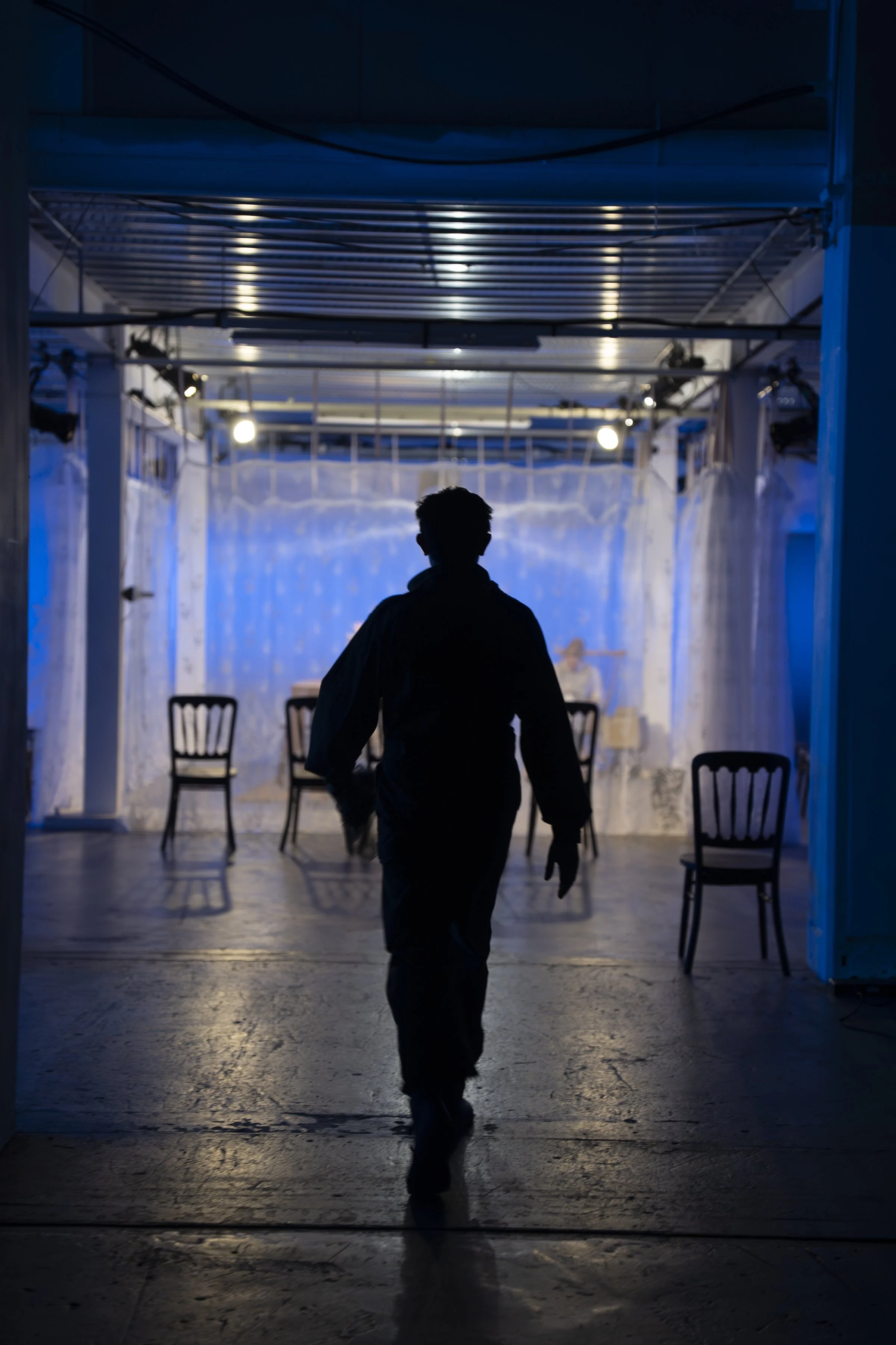 Silhouette of a person walking through a dimly lit space with chairs and a backdrop of a blue-lit curtain.