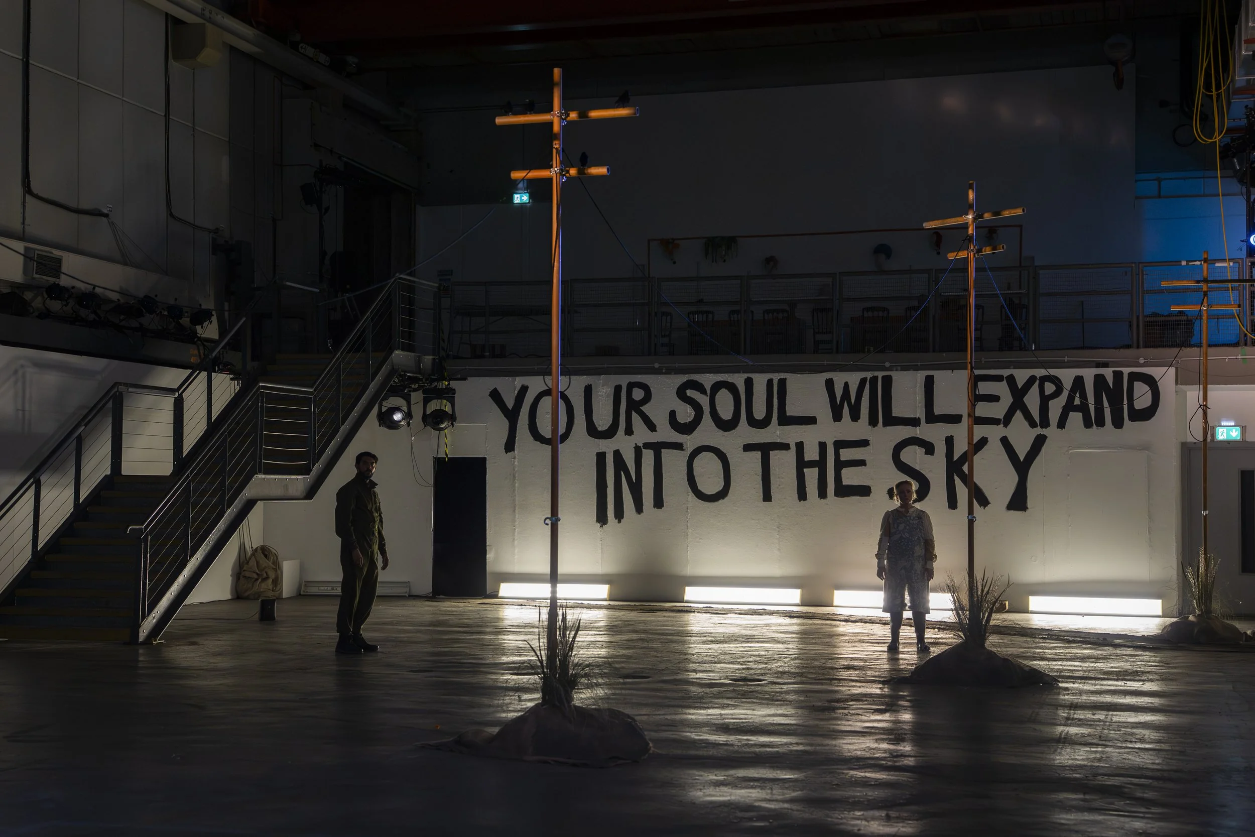 Interior of a building with a white wall, black painted letters saying "YOUR SOUL WILL EXPAND INTO THE SKY," and two people standing inside. There are wooden poles with wires, a staircase, and some plants on the floor.