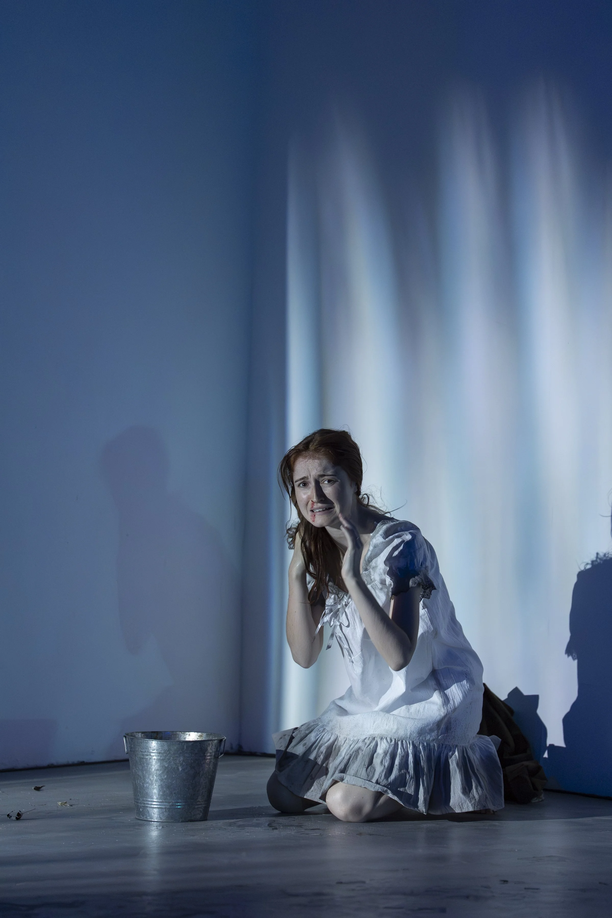 A woman with long reddish hair kneels on the floor, appearing distressed with a worried expression, in a dimly lit room with blue lighting. She wears a white dress with lace details and is reaching out with one hand. There's a metal bucket in front o