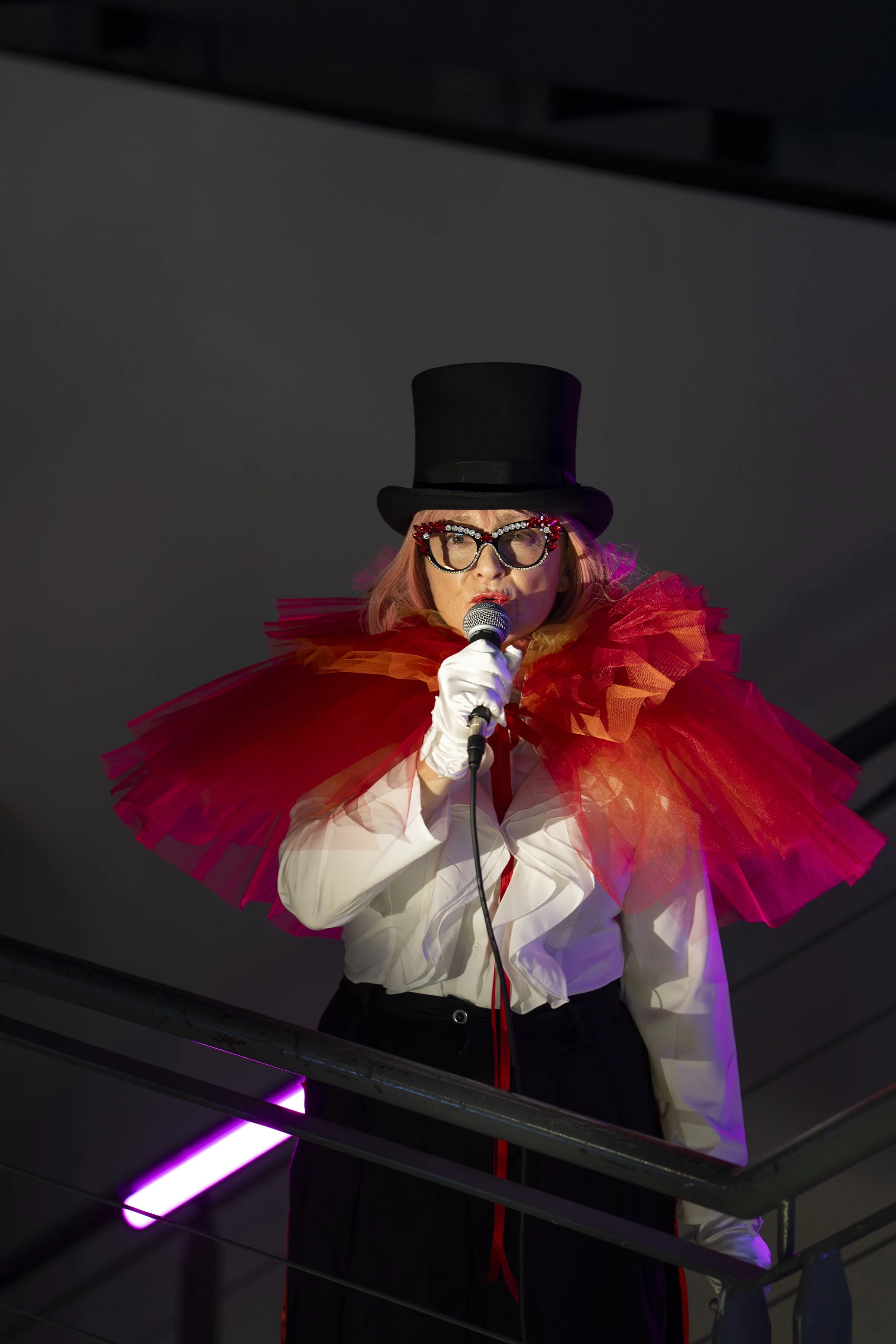 Person wearing a top hat, large glasses with red and clear embellishments, a white shirt, and a colorful tulle cape, singing into a microphone on a dimly lit stage.