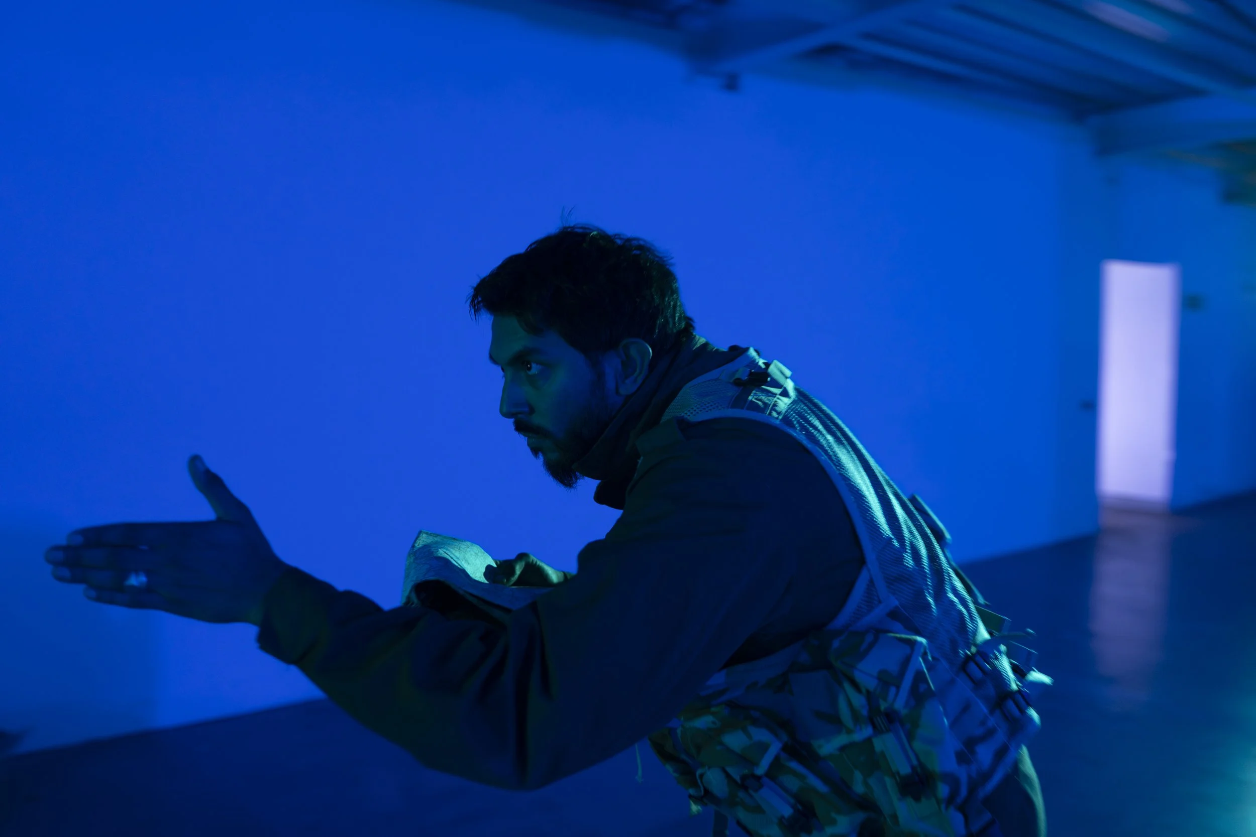 Man in camouflage jacket and backpack sitting in a room with blue lighting, looking at and using a smartphone.