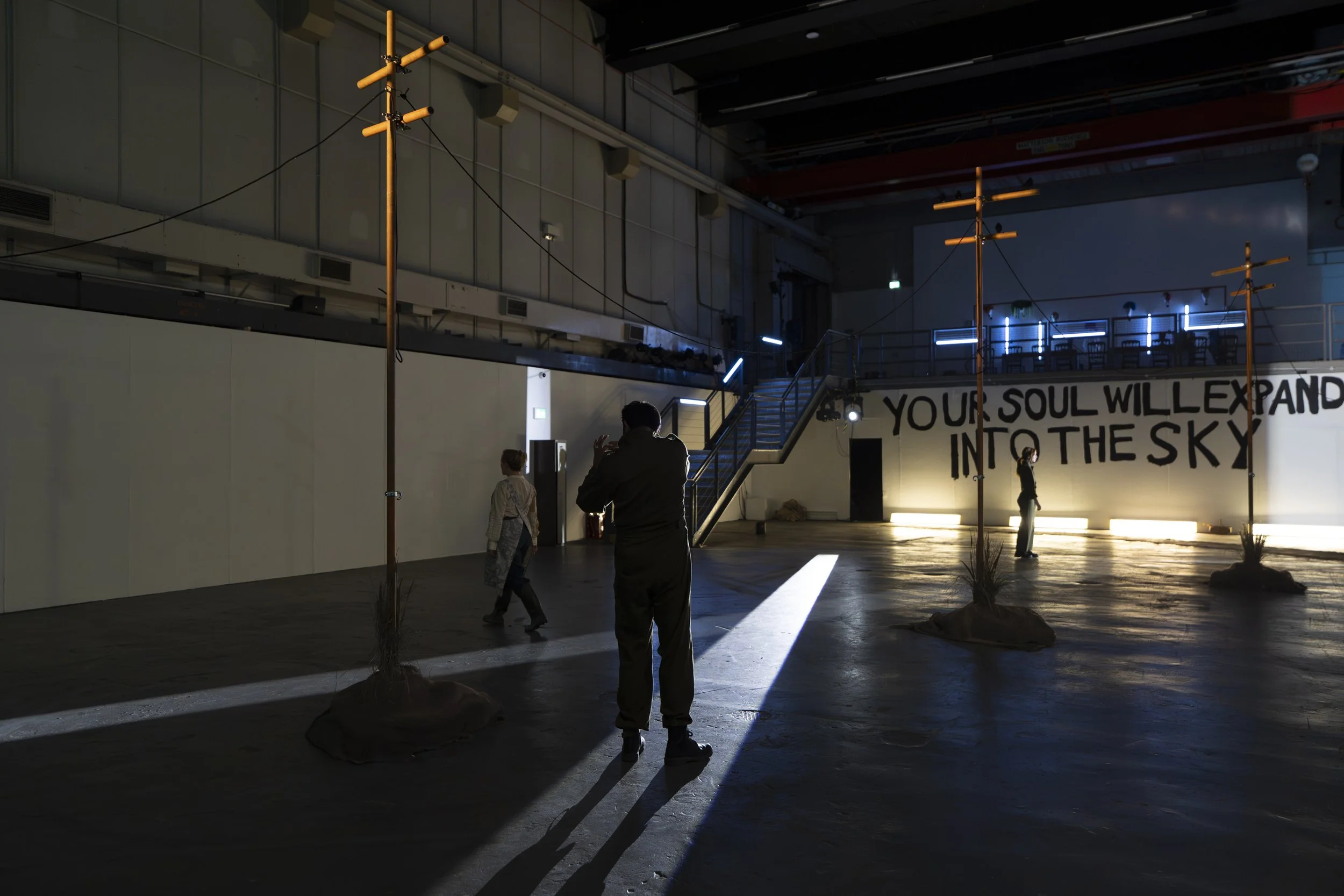 Dimly lit indoor space with several tall, thin poles with horizontal bars, some illuminated with neon lights. A staircase leading to an upper level, with people standing and walking around. A large message on the wall reads, 'YOUR SOUL WILL EXPAND IN