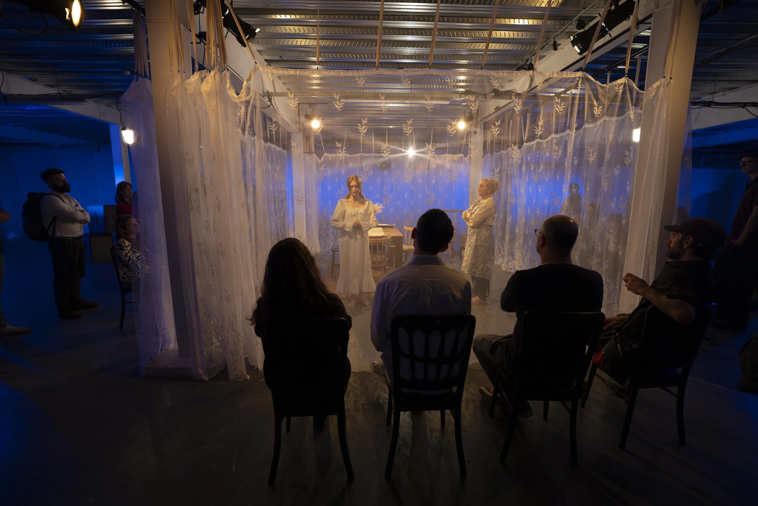 A theatrical performance or presentation taking place in a dimly lit room with an audience seated and watching two women on stage. The stage is decorated with sheer white curtains and soft, warm lighting, creating an intimate atmosphere.
