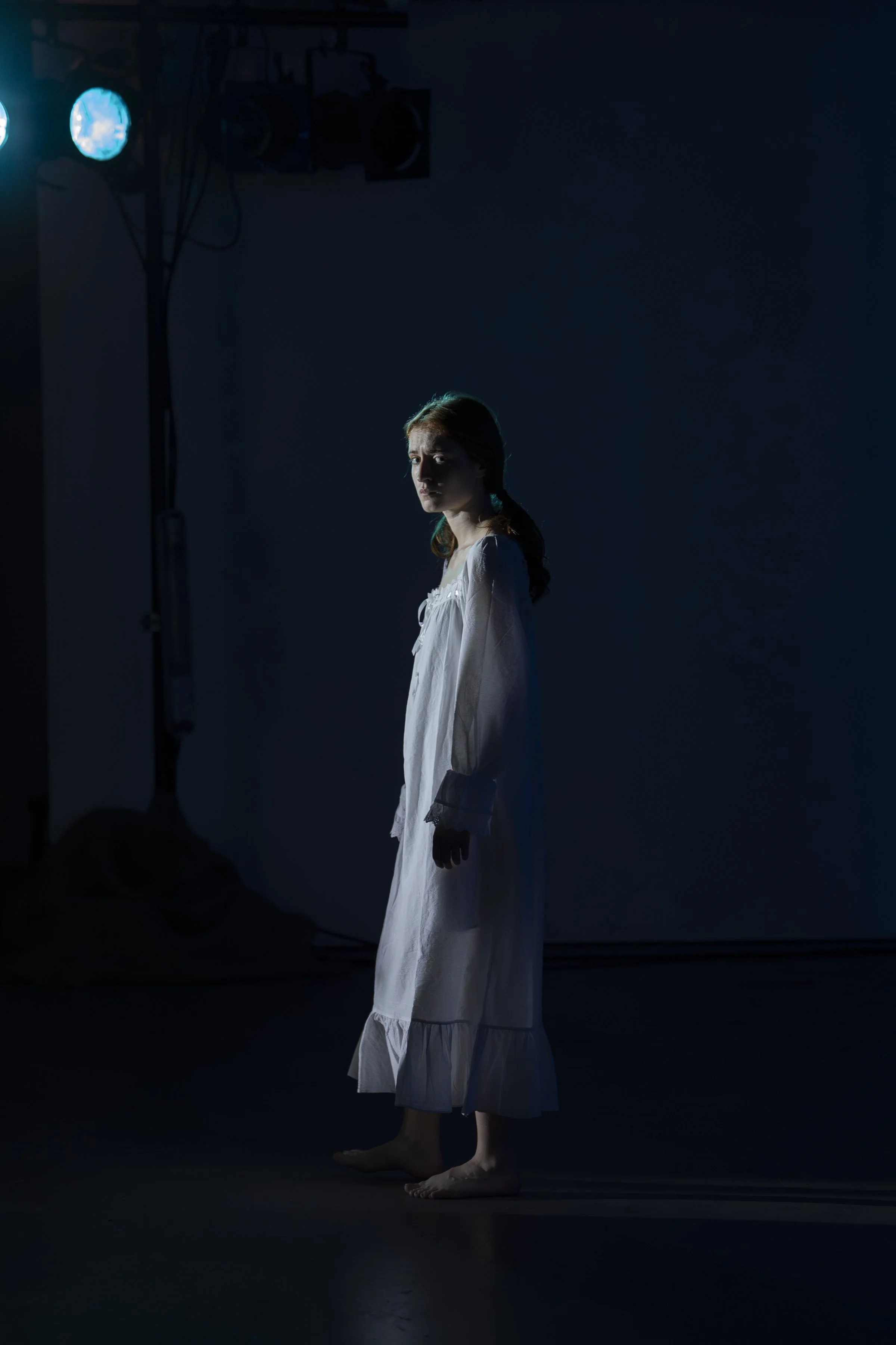 A young woman standing barefoot in a dark room with spotlight lighting, wearing a white nightgown, gazing thoughtfully.