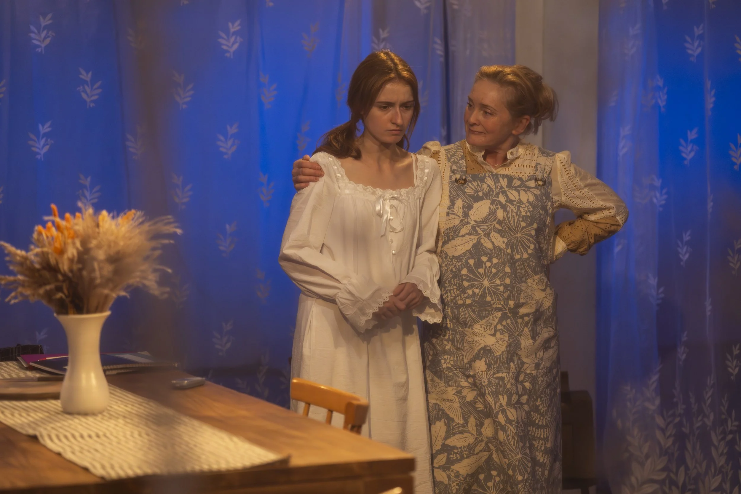 Two women, one young and one elderly, standing close together with the elderly woman’s arm around the younger woman’s shoulder, in a room with blue patterned curtains. A vase with dried flowers is on the dining table in the foreground.