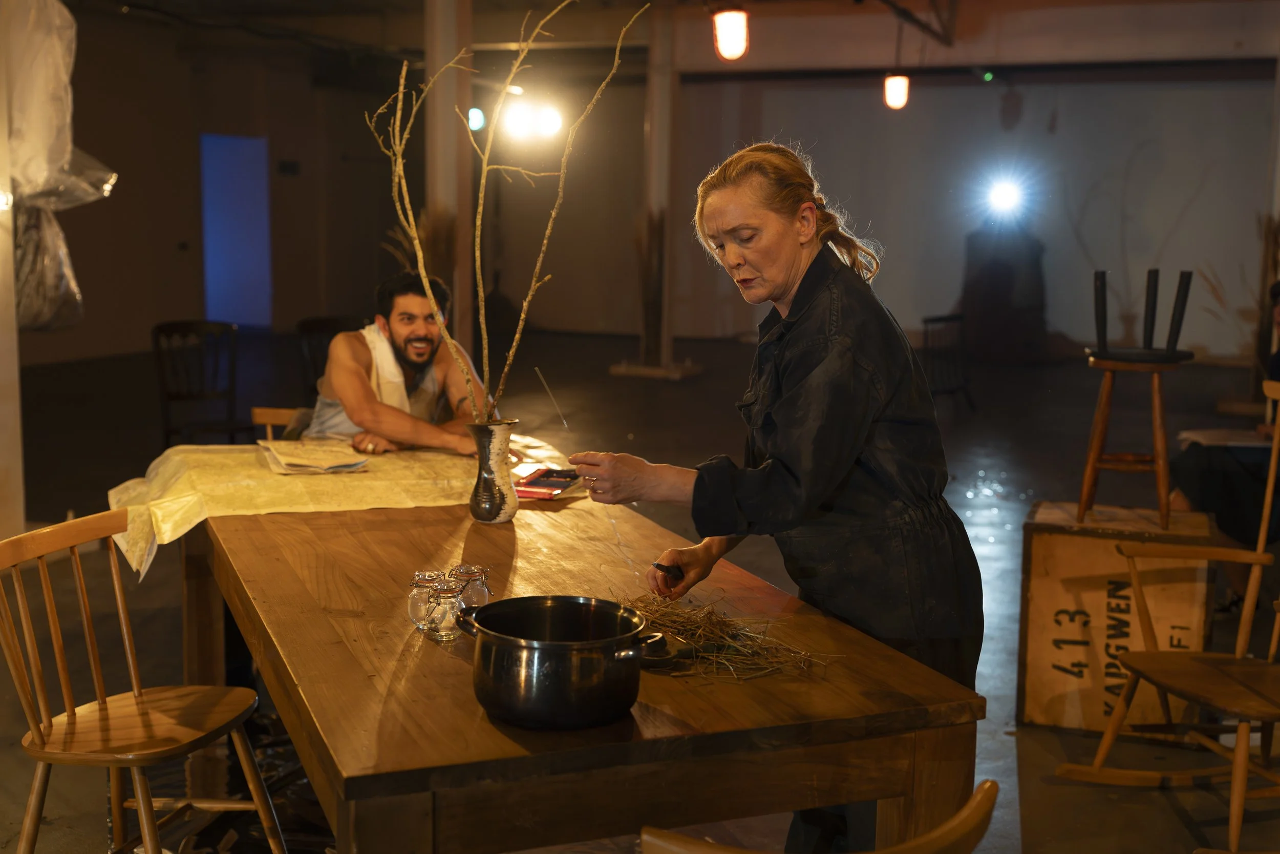 A woman with red hair standing at a wooden table preparing string, with a man smiling and looking on in the background. The environment appears to be a dimly lit art studio or workshop with chairs and boxes around.