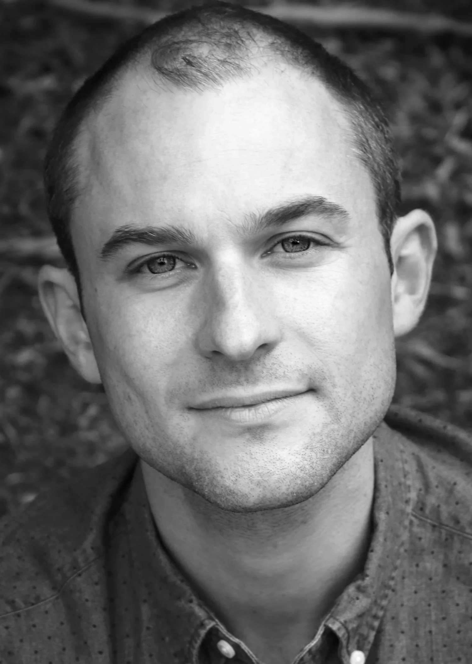 Black-and-white close-up photograph of a young man's face, showing only the upper part, including his eyes, forehead, and part of his mouth. He has light skin, short dark hair, and is wearing a collared shirt. The background is out of focus and appears to be outdoor ground or dirt.