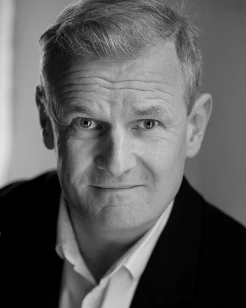 Black and white close-up portrait of a middle-aged man with short, light hair, wearing a collared shirt and blazer.
