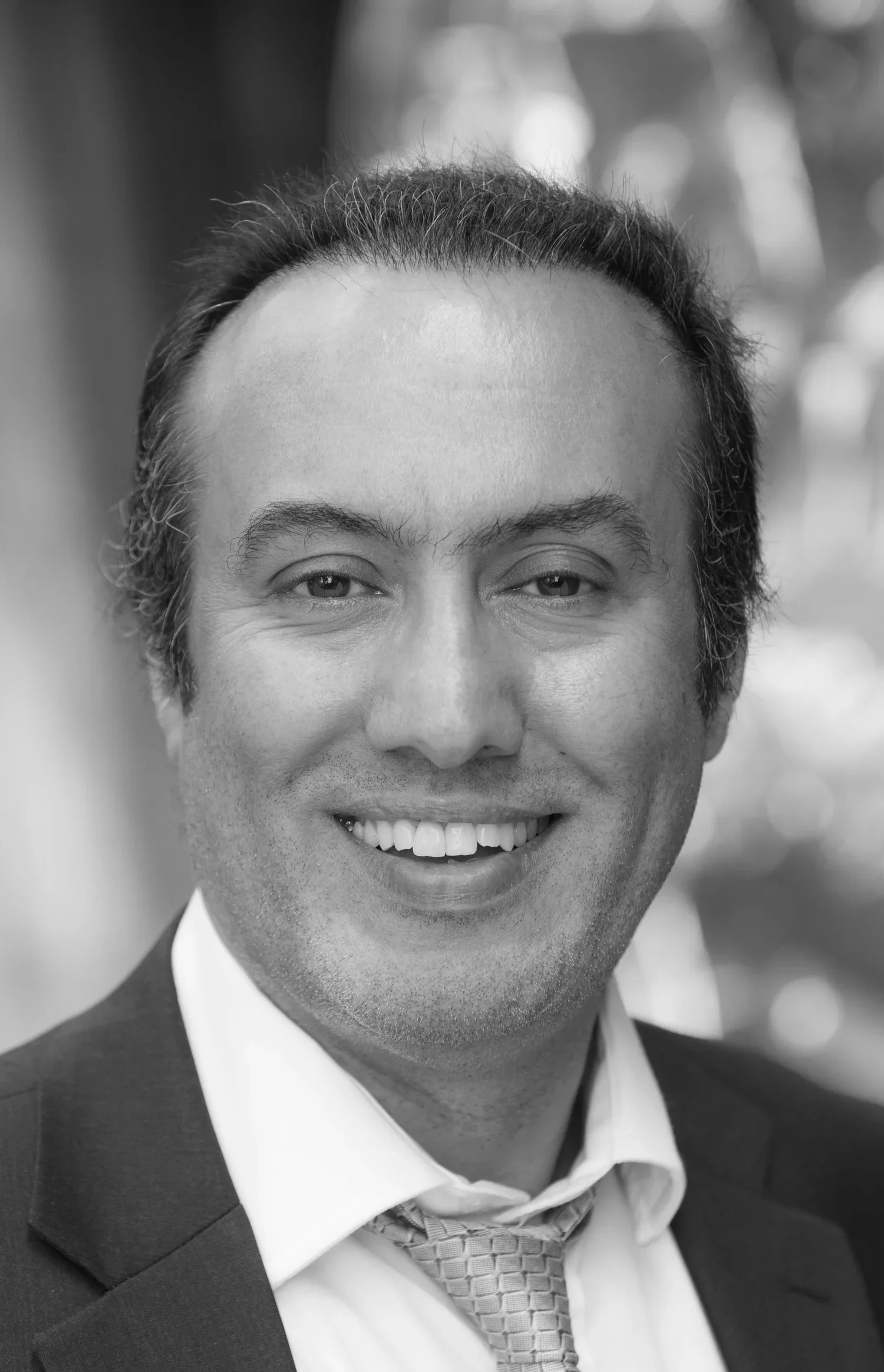 Black and white photograph of a man smiling in a business suit, white shirt, and patterned tie, with a blurred background.