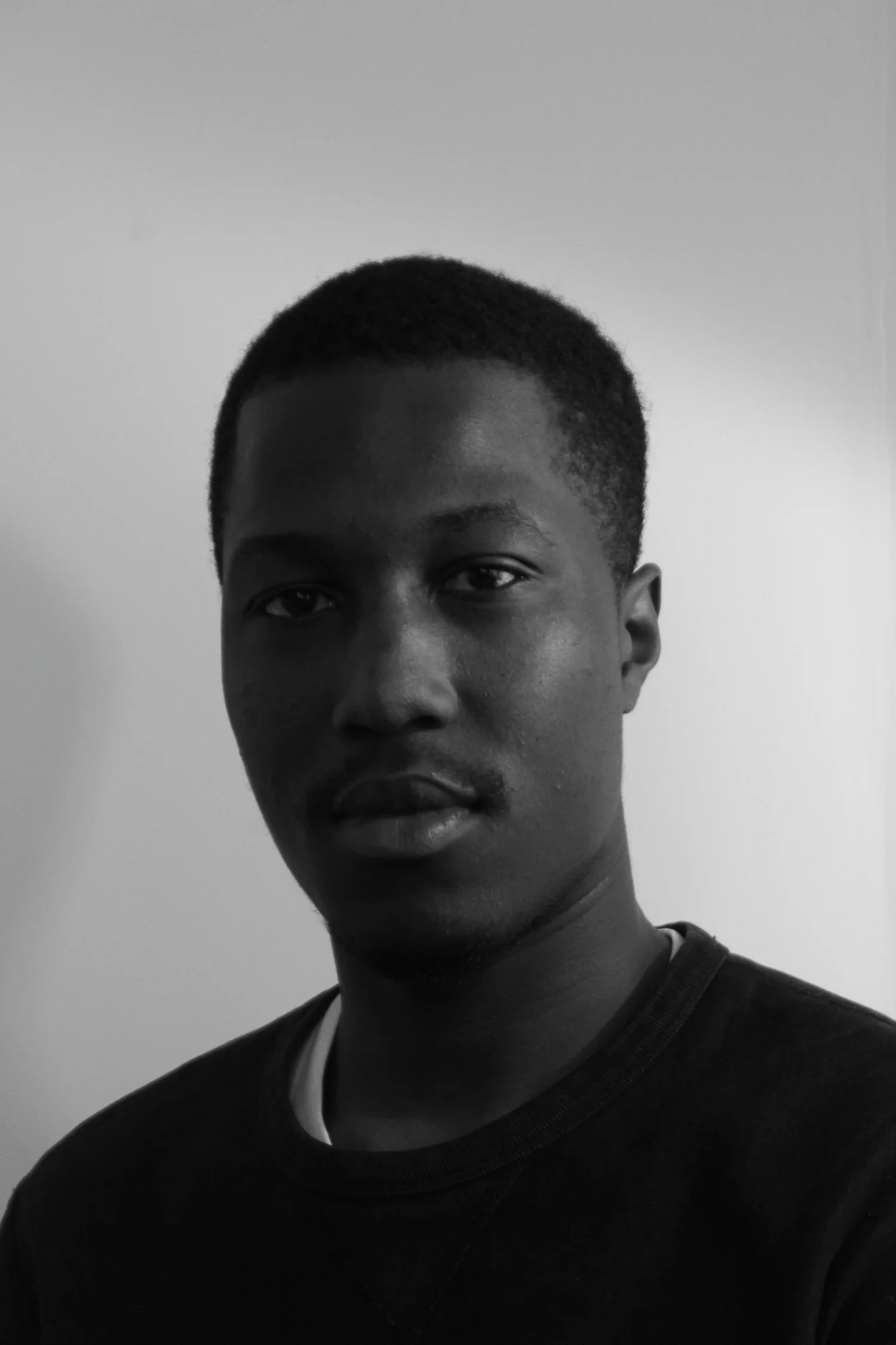Black and white photo of a young man with short hair, looking at the camera, wearing a black shirt with a white collar, against a plain background.