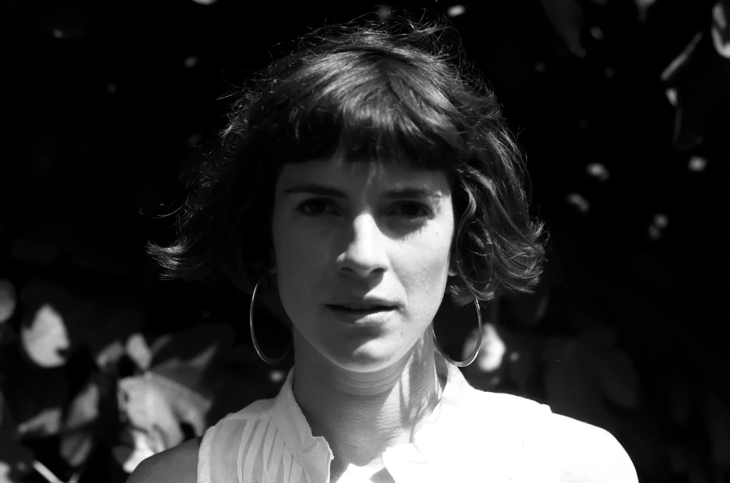 A black and white photo of a woman with short curly hair, wearing large hoop earrings and a collared shirt, standing outdoors with blurred foliage in the background.