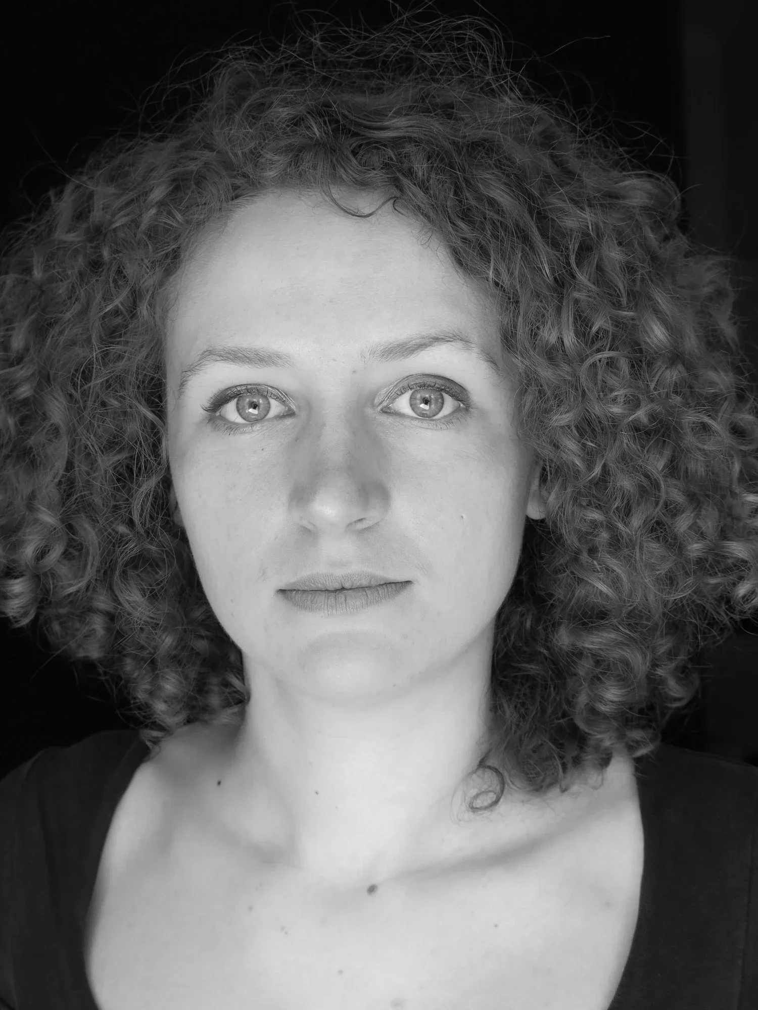 Black and white portrait of a woman with curly hair, visible eyes, and part of her face, with a dark background.