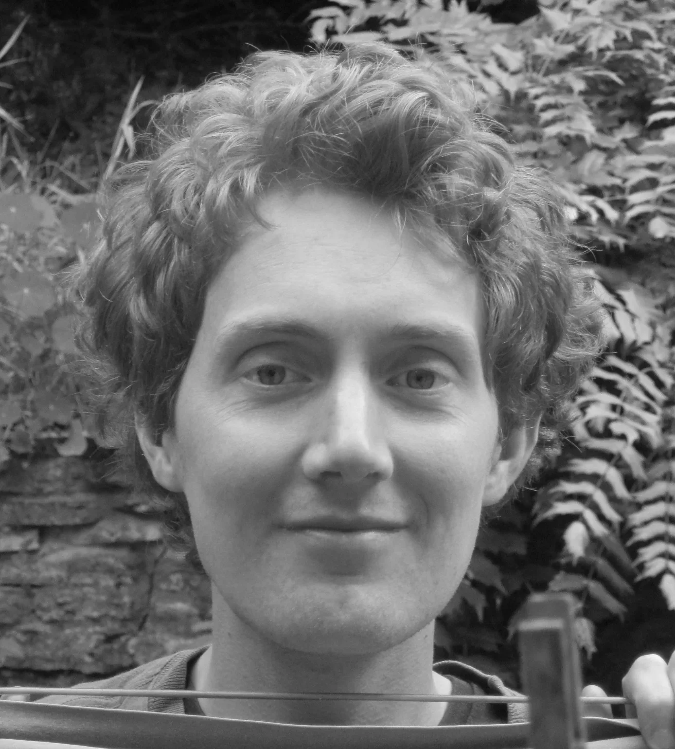 Black and white photograph of a person with curly hair, looking directly at the camera, with a background of plants and foliage.