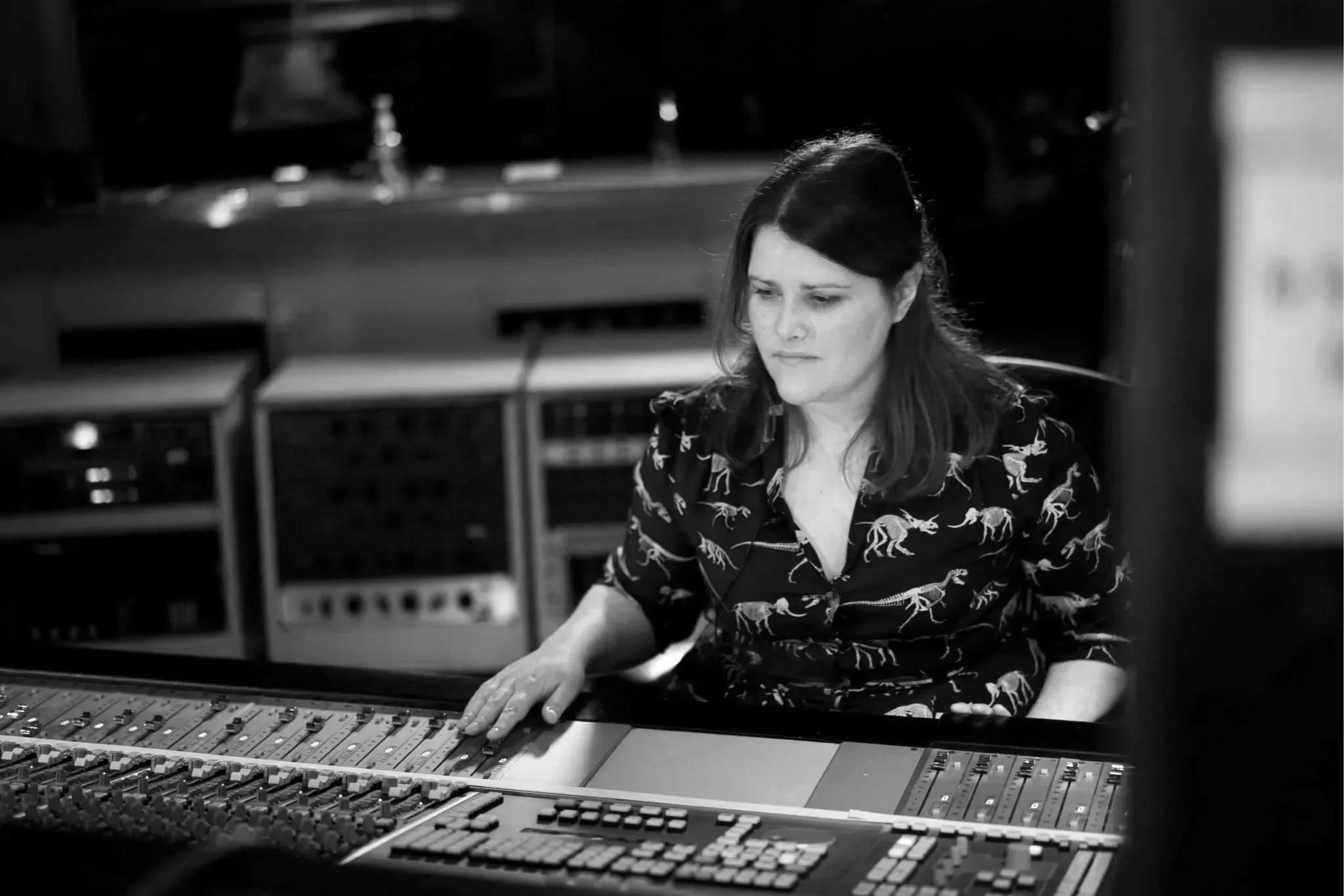 Black and white photograph of a woman with shoulder-length dark hair working with audio mixing equipment in a recording studio or sound booth.