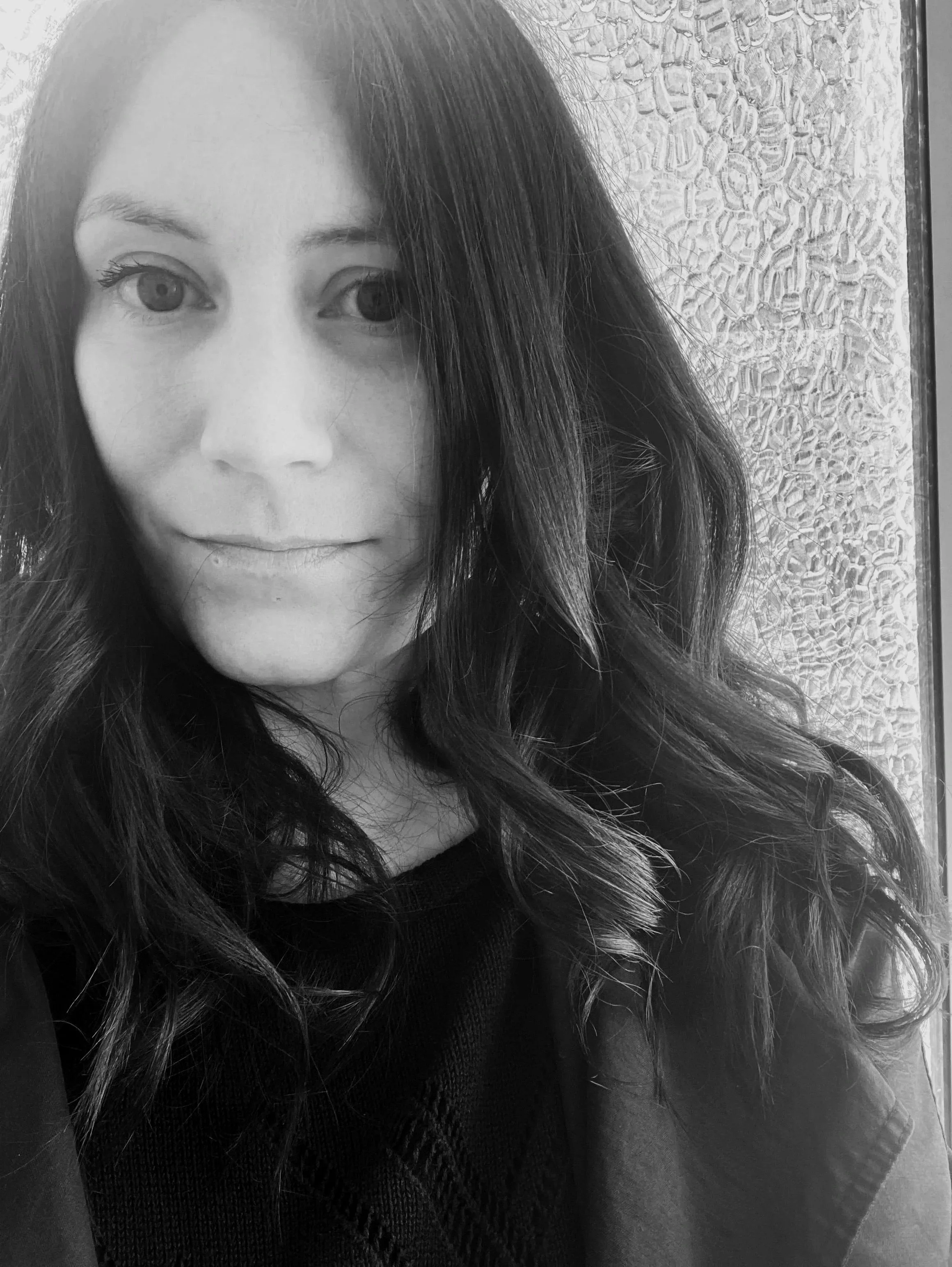 A close-up black and white photograph of a woman with long, wavy hair, looking at the camera, with a textured wall in the background.