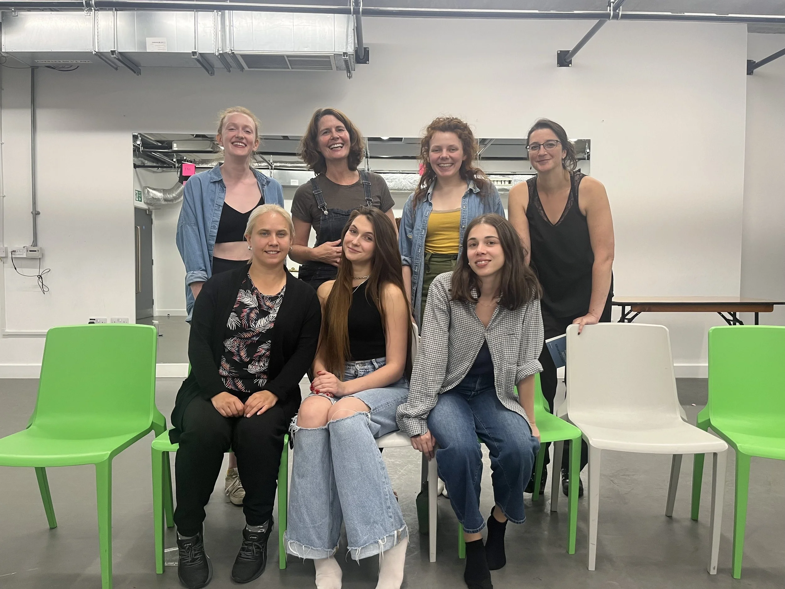 Group of seven women posing and smiling for a photo in an indoor space with white walls and green and white chairs.