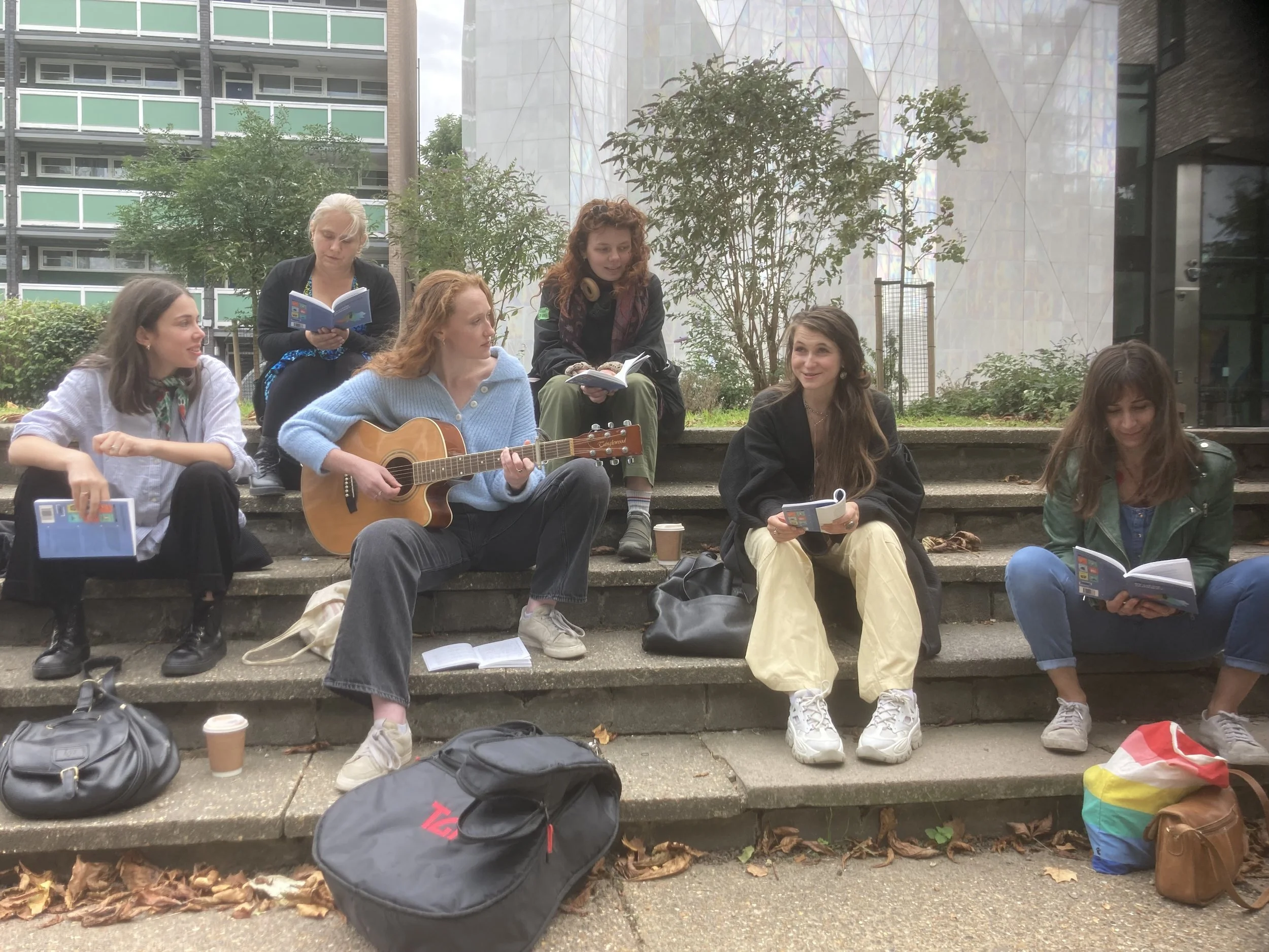 A group of women sitting on steps outdoors, some reading and one playing guitar, with backpacks and coffee cups around them, in an urban setting with modern buildings and trees in the background.