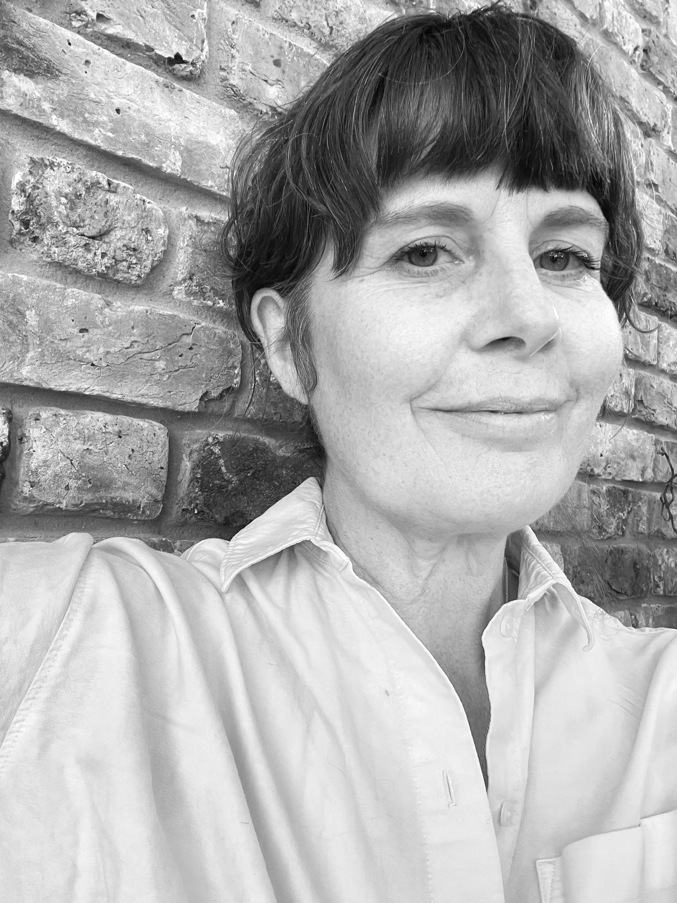 A black and white close-up portrait of a woman with short hair, wearing a button-up shirt, standing in front of a brick wall.