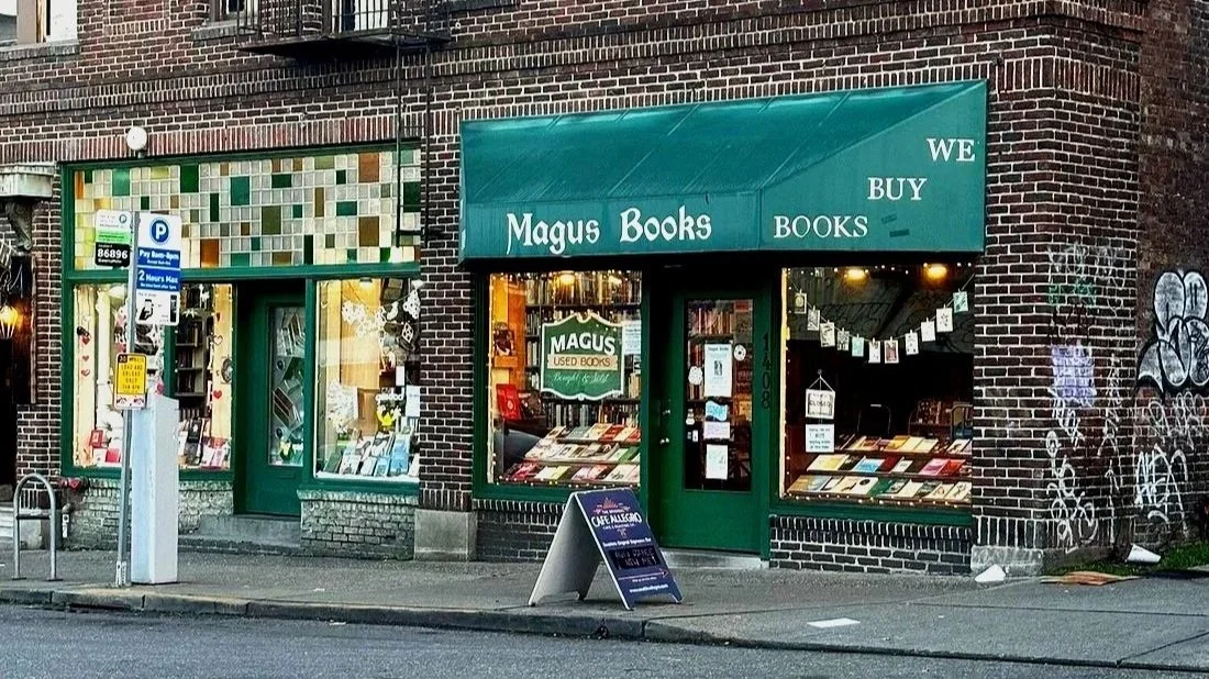 A bookstore storefront with a green awning that reads "Magus Books" and "WE BUY BOOKS." The shop has large display windows showcasing books and a sidewalk chalkboard sign outside.
