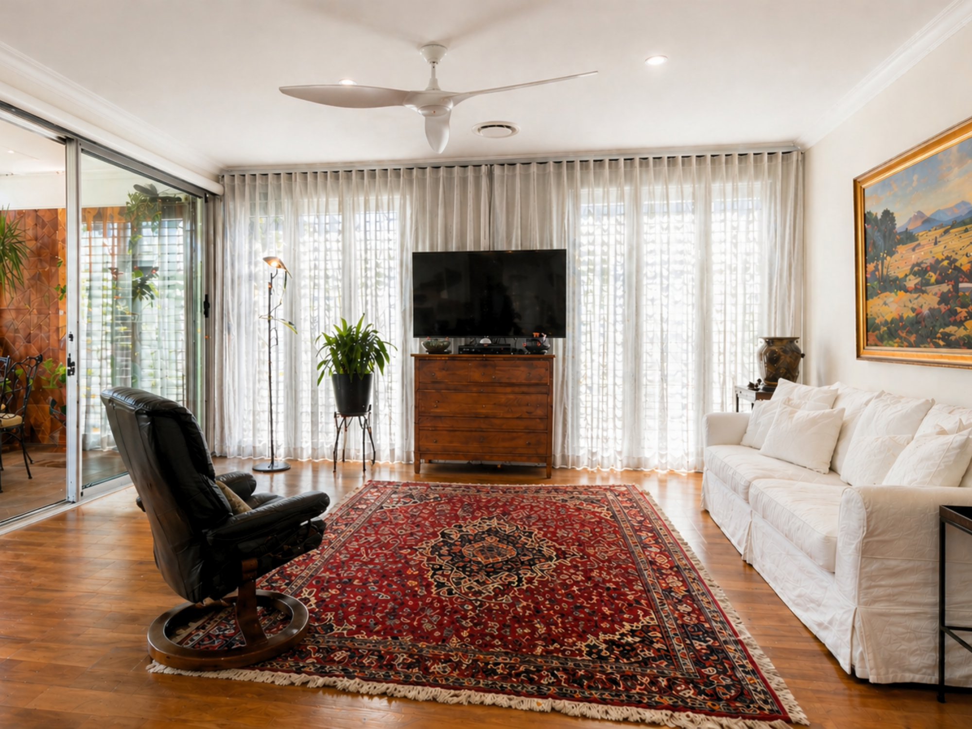 Greenville, South Carolin living room with hardwood floors, a large red patterned area rug, white curtains covering windows, a flat-screen TV on a wooden stand, a black recliner, a white sofa, and a ceiling fan.