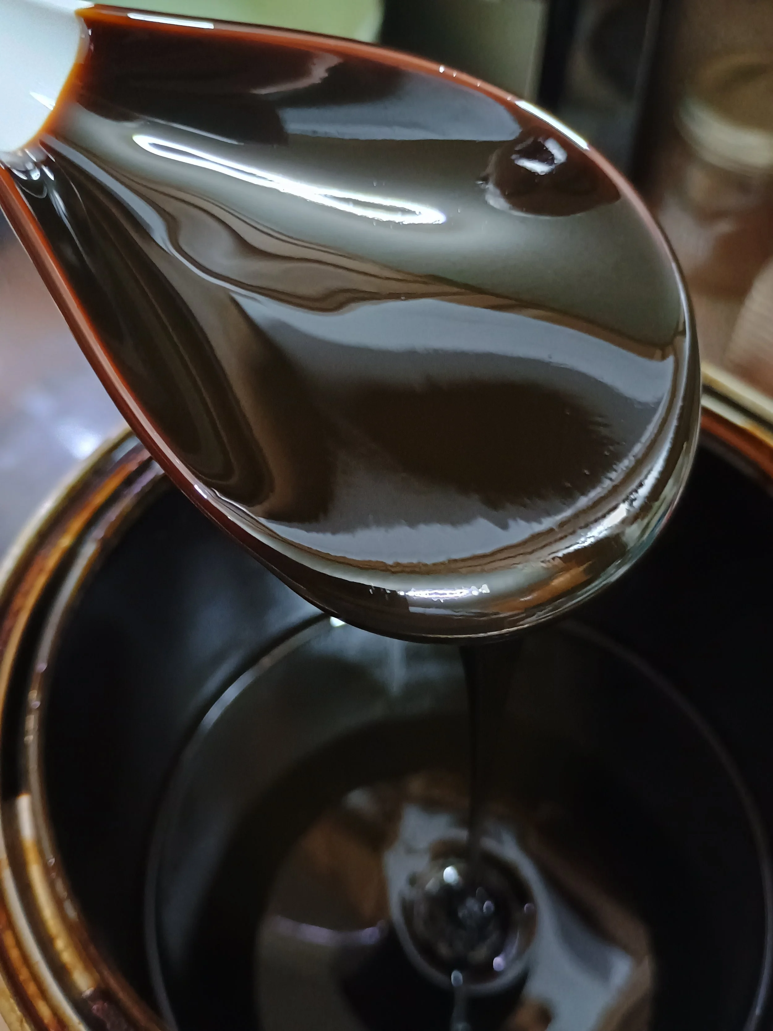 A bird's-eye view of a white plastic spoon drizzling down black pine tar into a metal can