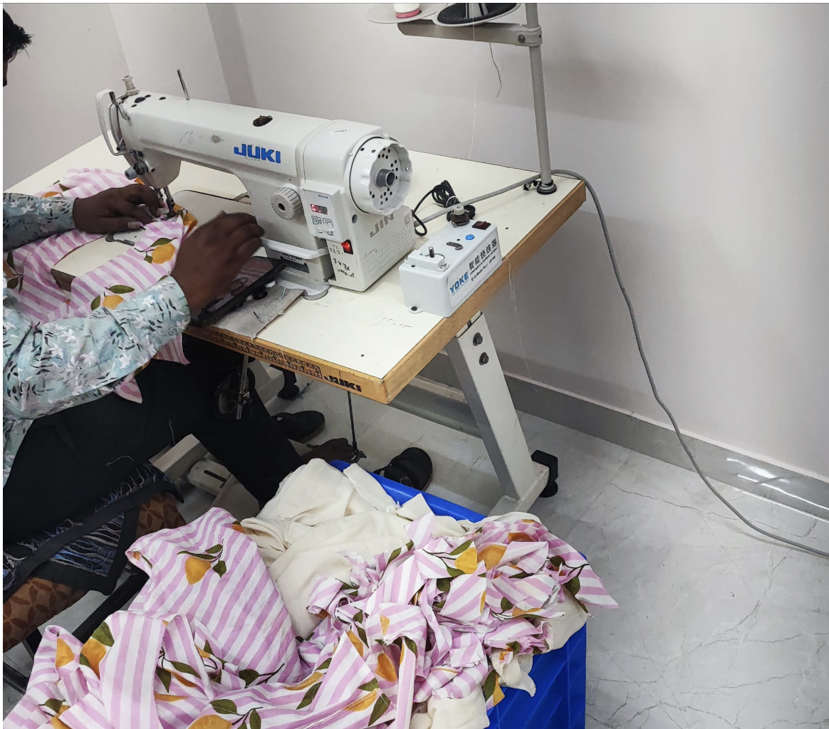 Une personne utilisant une machine à coudre industrielle blanche avec le mot 'JUKI' bleu, située sur un bureau en bois blanc, en train de coudre un tissu à rayures roses, blanches et décorations de fruits orange. Un bac en plastique bleu contenant des morceaux de tissu est placé sous le bureau.