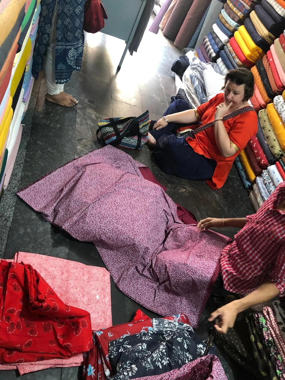 Une femme assise à terre dans un magasin de tissus, entourée de rouleaux de tissus colorés empilés. Elle porte un vêtement rouge et regarde pensivement. Une autre personne est partiellement visible, tenant un tissu rose à motifs. Des sacs et autres tissus sont également visibles dans l'image.