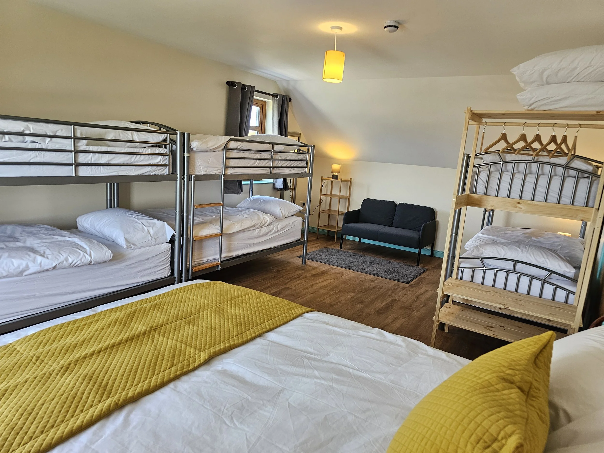 A bedroom with multiple bunk beds, a small couch, a window with dark curtains, a lamp, and a ceiling light in an attic space with sloped ceilings.
