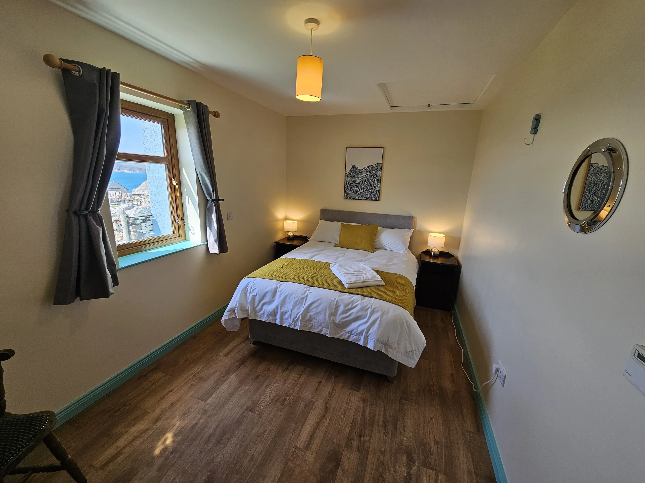 A cozy bedroom with a bed, yellow bedding, and a yellow pillow by a window with dark curtains. There are two nightstands with lamps, a framed picture above the bed, a circular mirror on the wall, and hardwood floors.