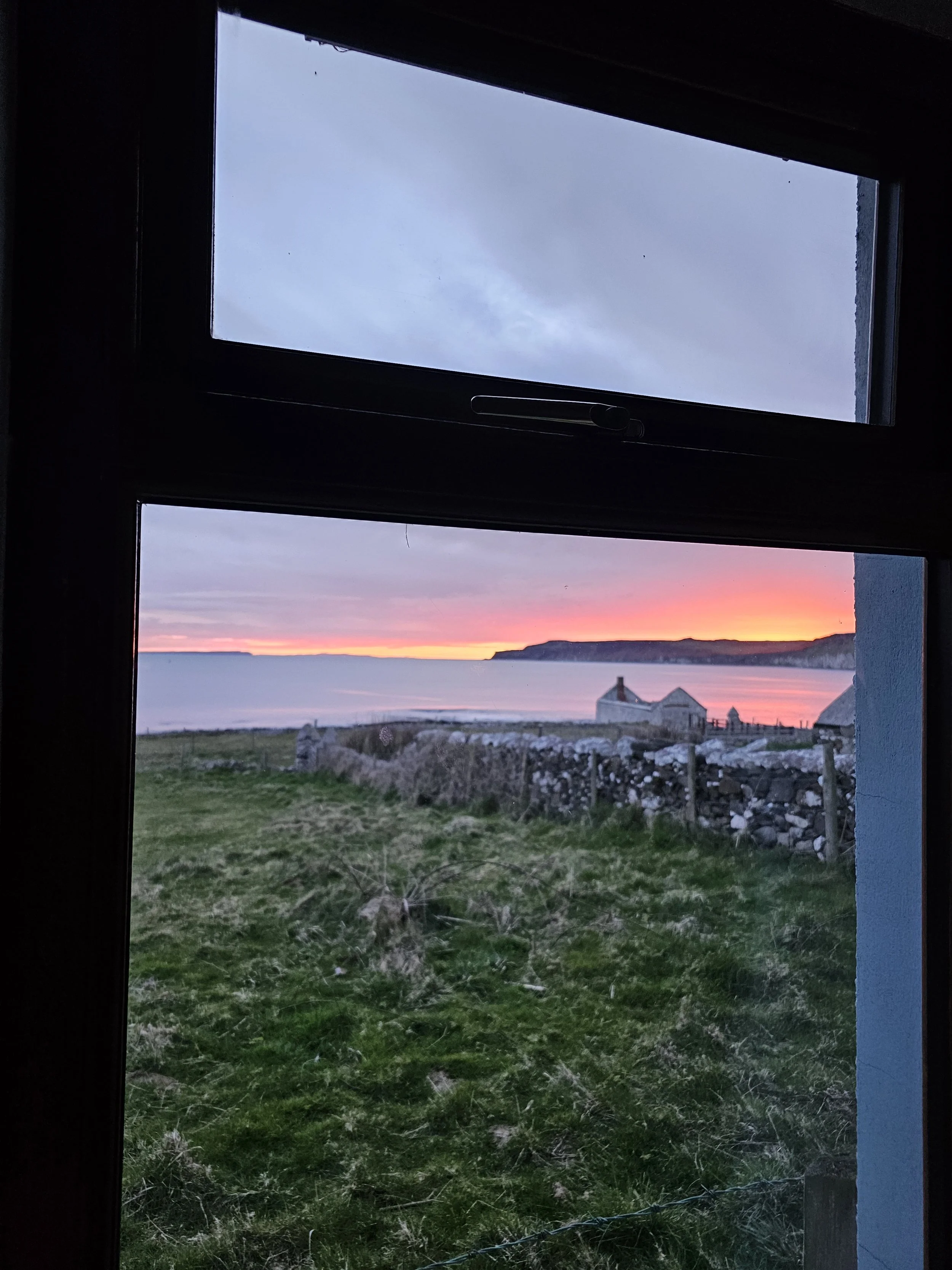 View of a sunset over a coastal landscape seen through a window, with grassy foreground, stone wall, and houses by the shore.