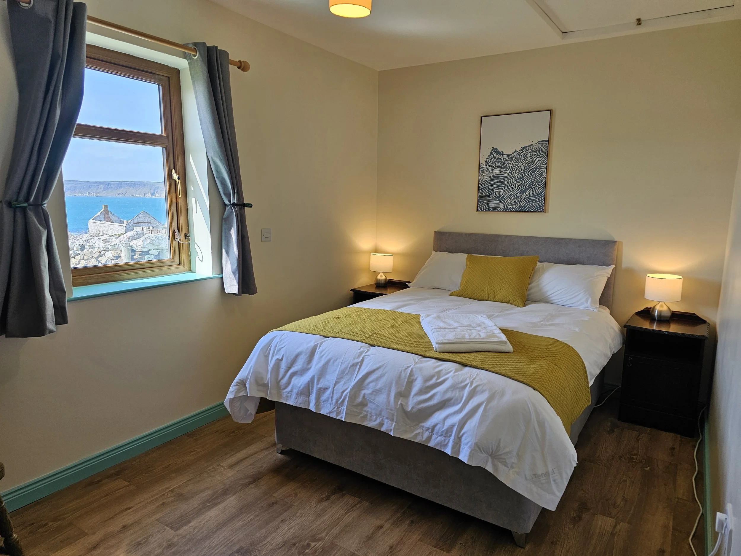 A cozy bedroom with a large bed covered in white bedding and a yellow quilted runner. There are two bedside tables each with a small lampshade. The room has a window with dark curtains, and outside the window, there is a view of houses and a body of water with a coastline in the background. A piece of wall art featuring a wave is hung above the bed.